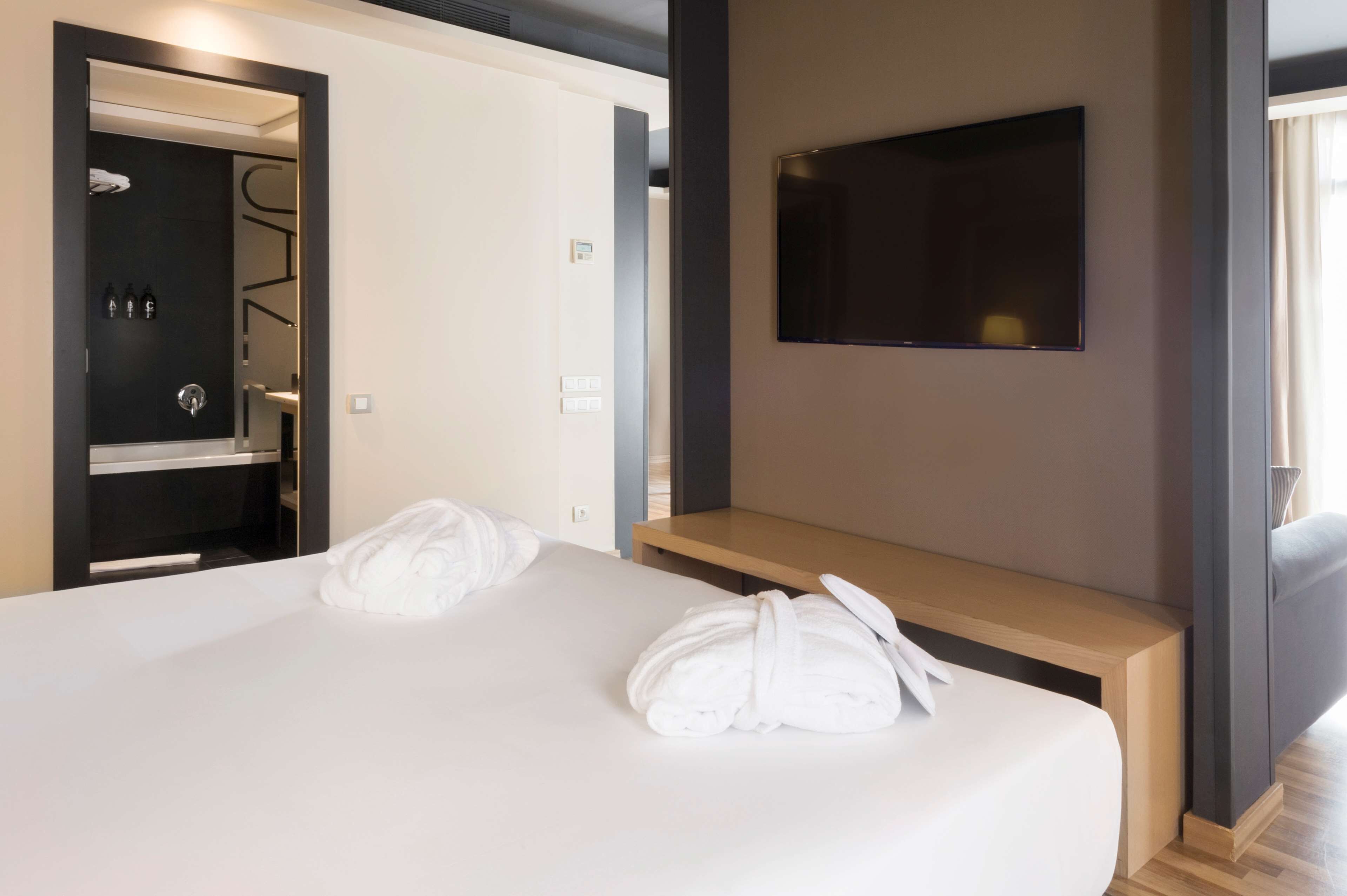 A hotel room with a bed, a TV mounted on the wall, and a wooden bench.
