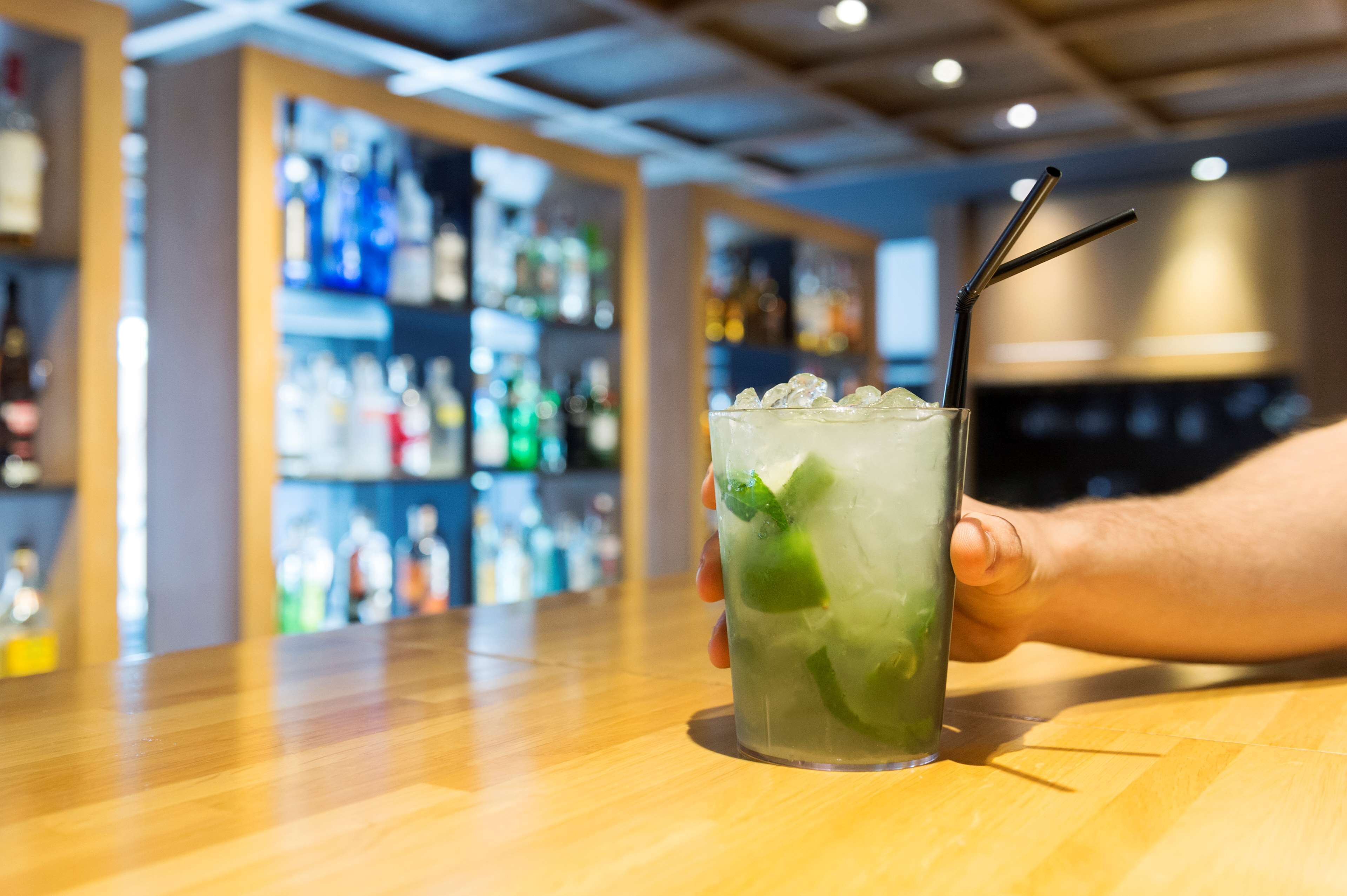 A bar setting with a hand holding a glass of a green cocktail with ice and a straw.