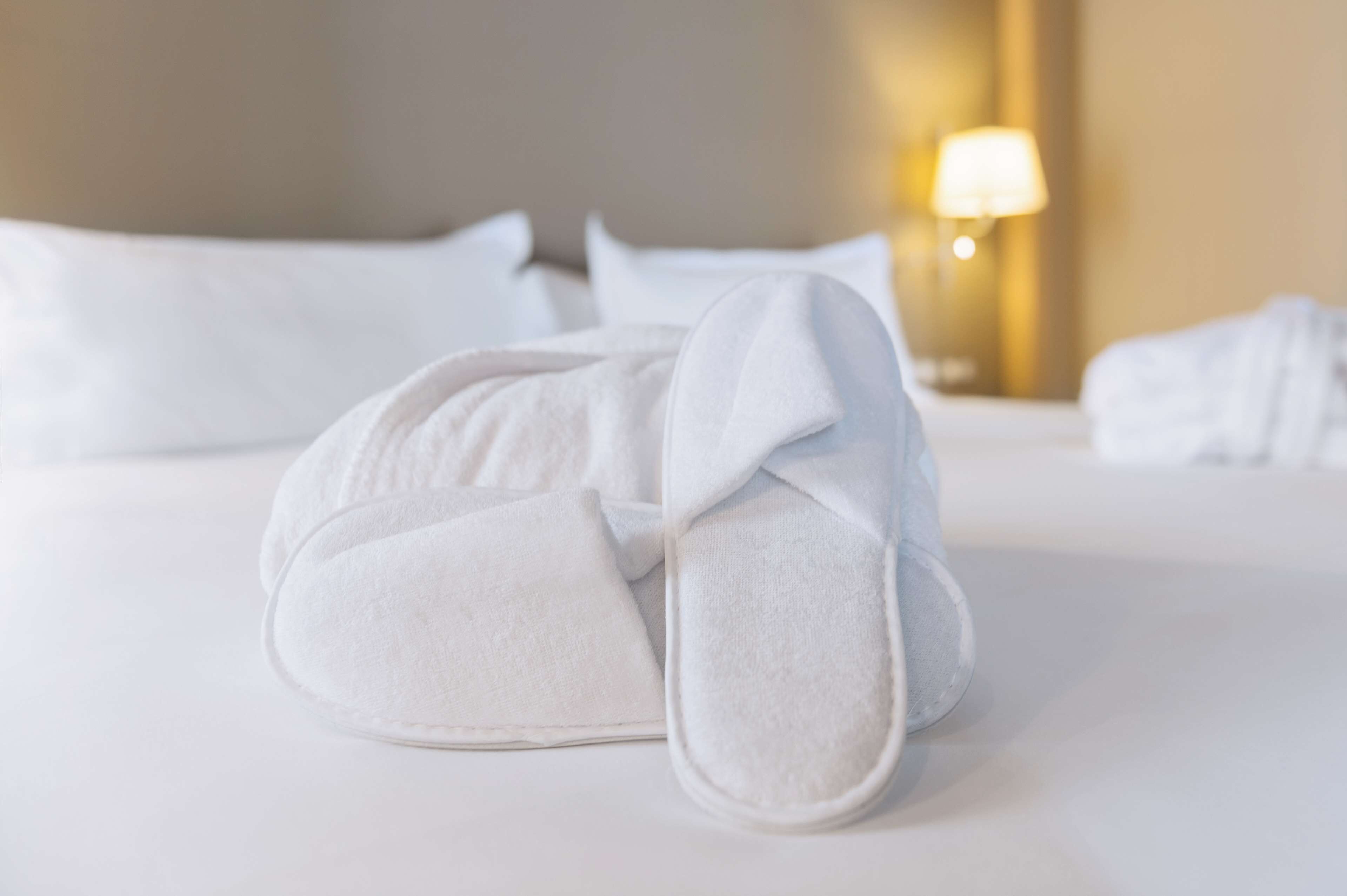 Toalla blanca bien doblada y pantuflas sobre una cama con ropa de cama y almohadas blancas.