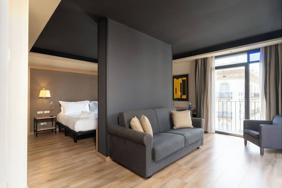 Una habitación de hotel moderna con un sofá gris, una cama con sábanas blancas, una mesita de noche pequeña con una lámpara y vistas a la ciudad a través de amplias ventanas.