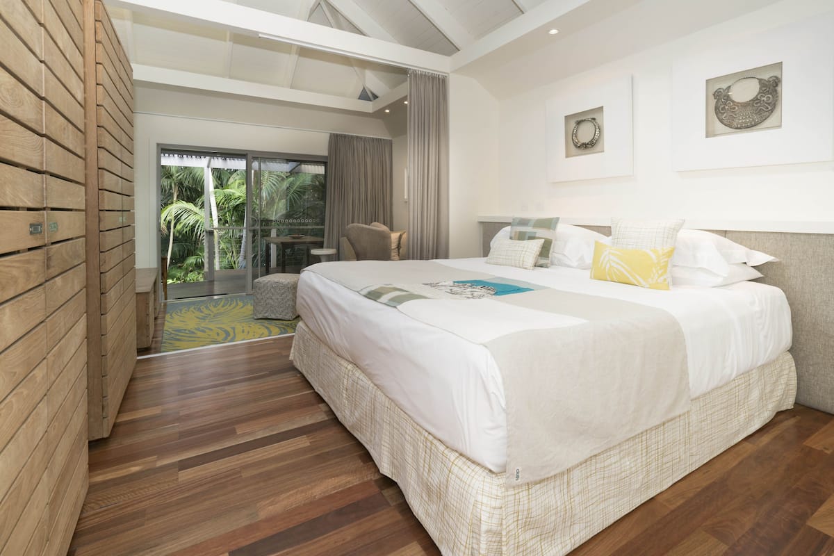 Un espacioso dormitorio con una cama grande, un balcón con vistas a palmeras y un diseño moderno con suelo de madera y techo de cristal.