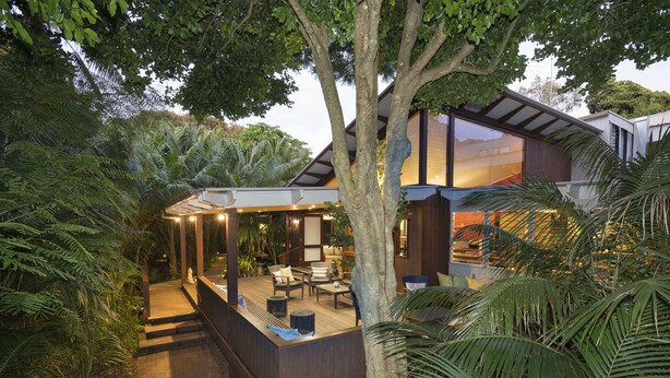 Une maison moderne dotée d’une terrasse en bois, entourée d’une végétation luxuriante et de palmiers.