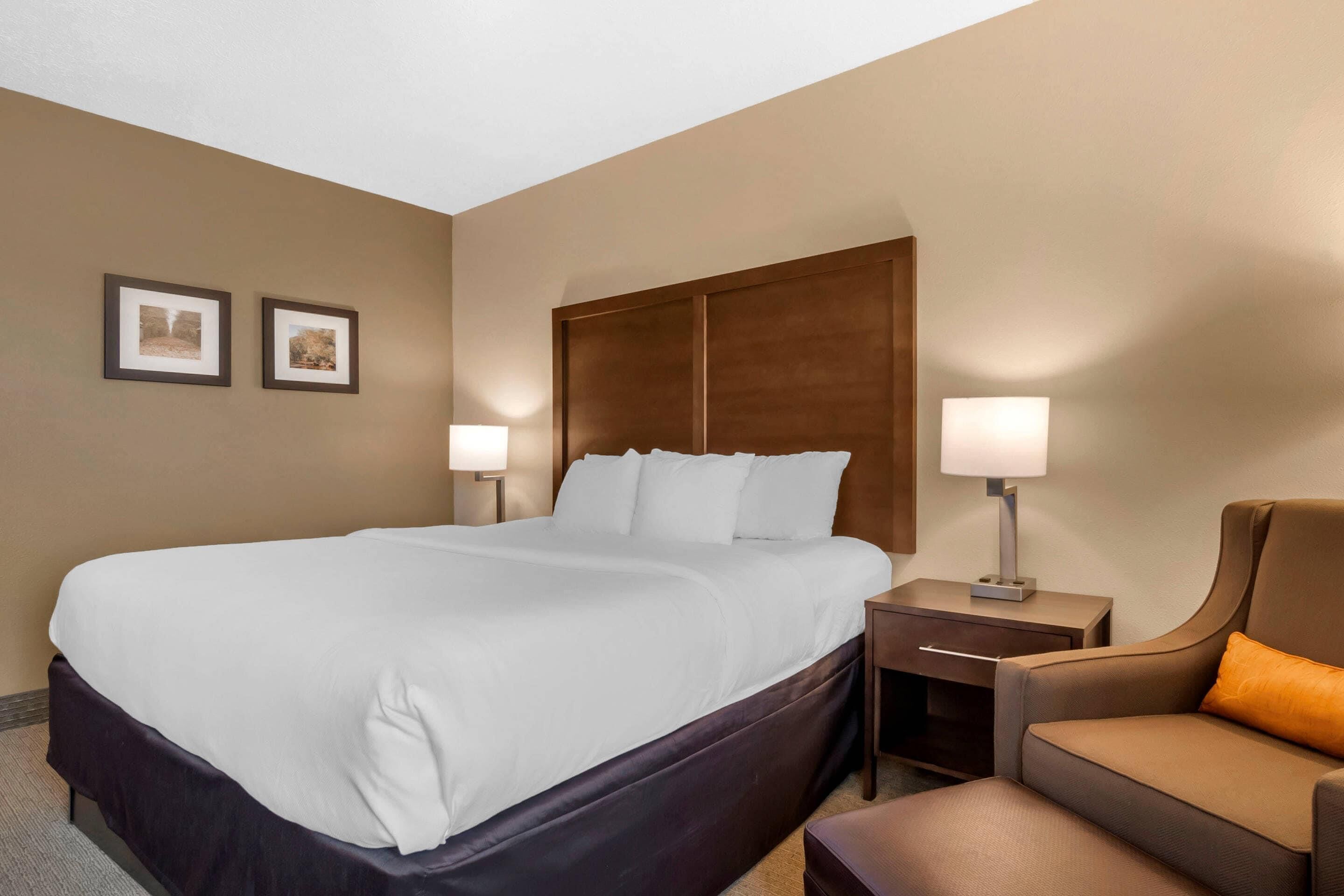Une chambre d’hôtel avec un grand lit, deux tables de chevet avec des lampes, une tête de lit en bois et un fauteuil brun.