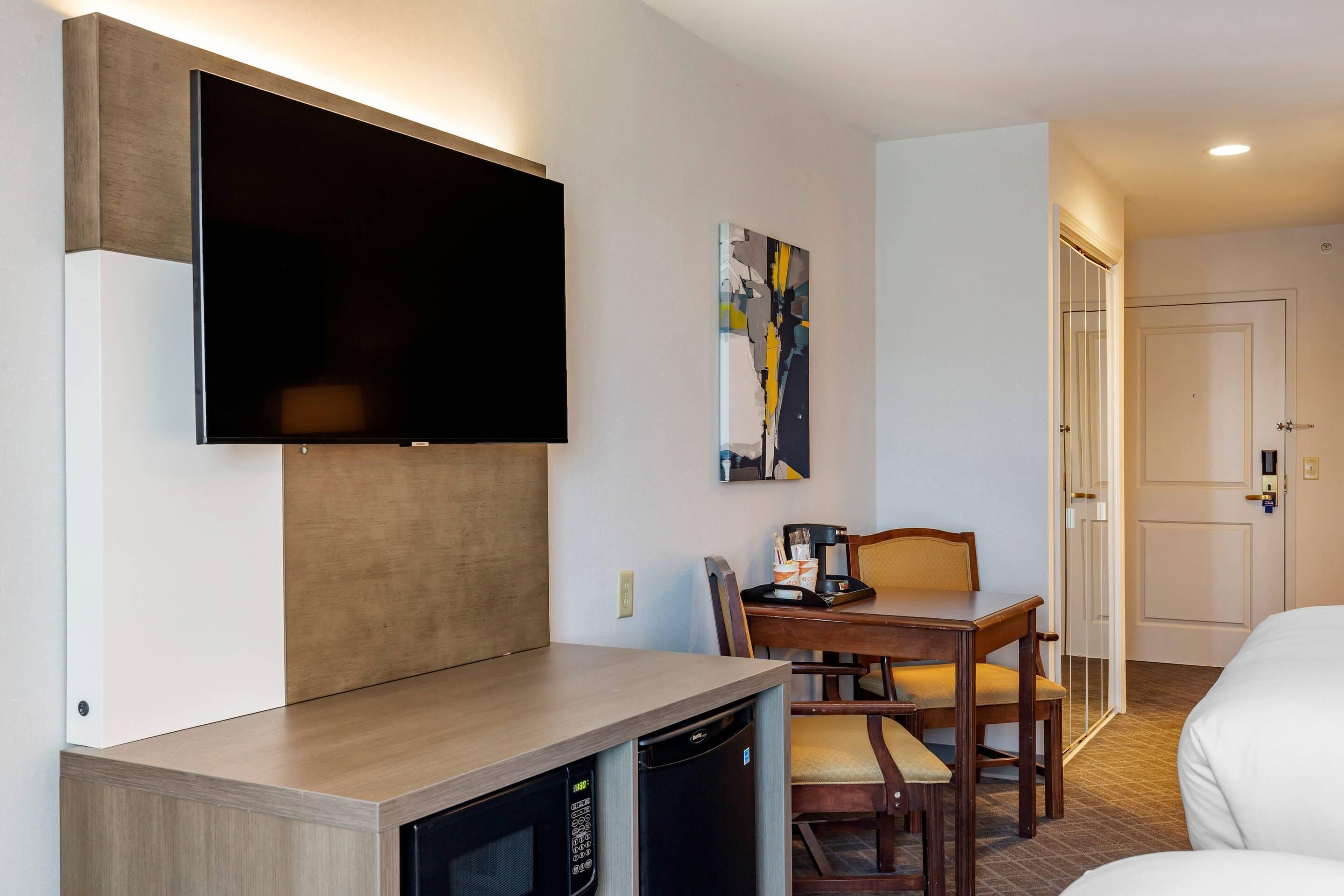 A hotel room with a flat-screen TV mounted on the wall, a small dining table with chairs, a coffee maker, and a bed.