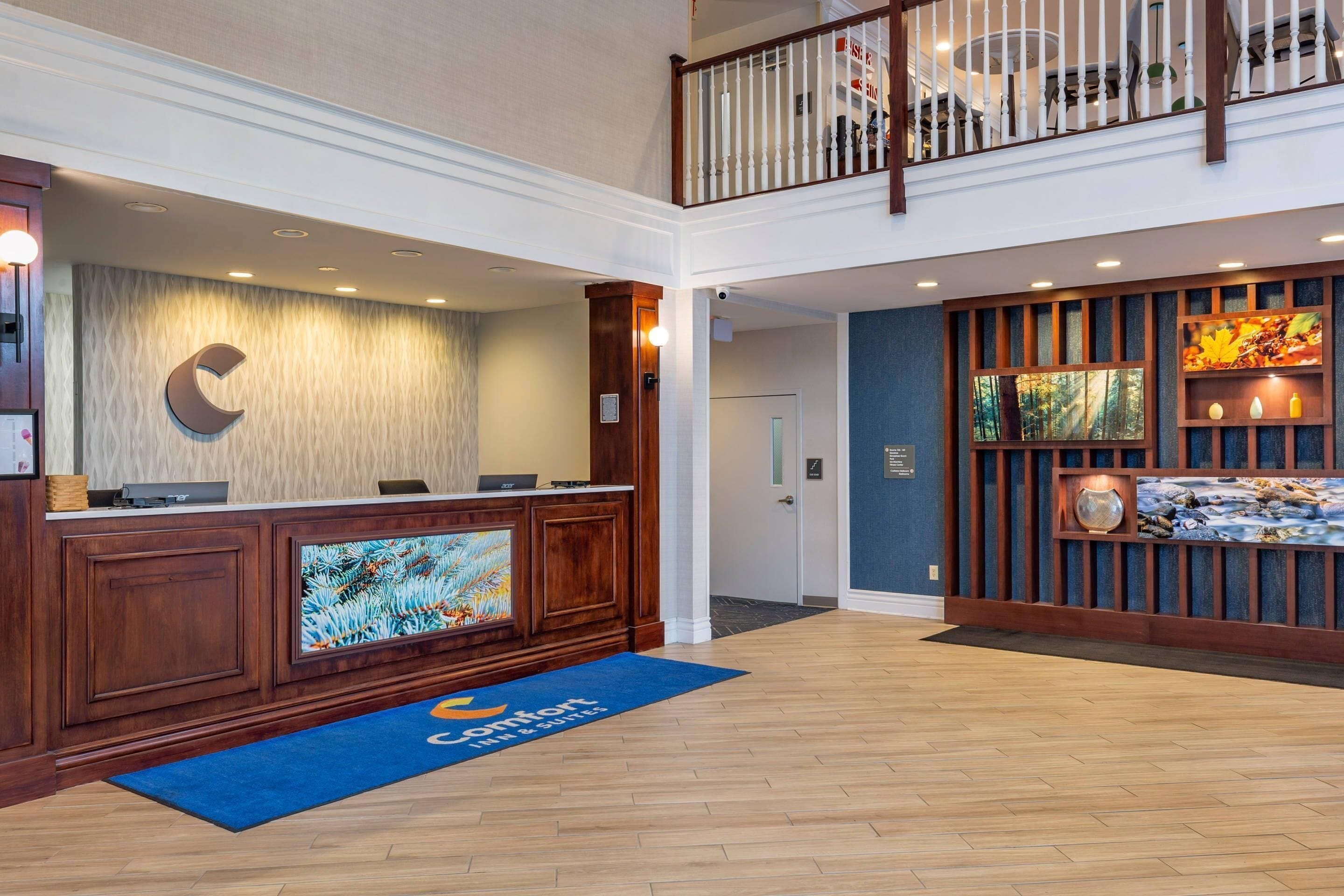 A hotel reception area with a wooden reception desk, a blue carpet with the hotel logo, a large C-shaped logo on the wall, and a balcony with a railing.
