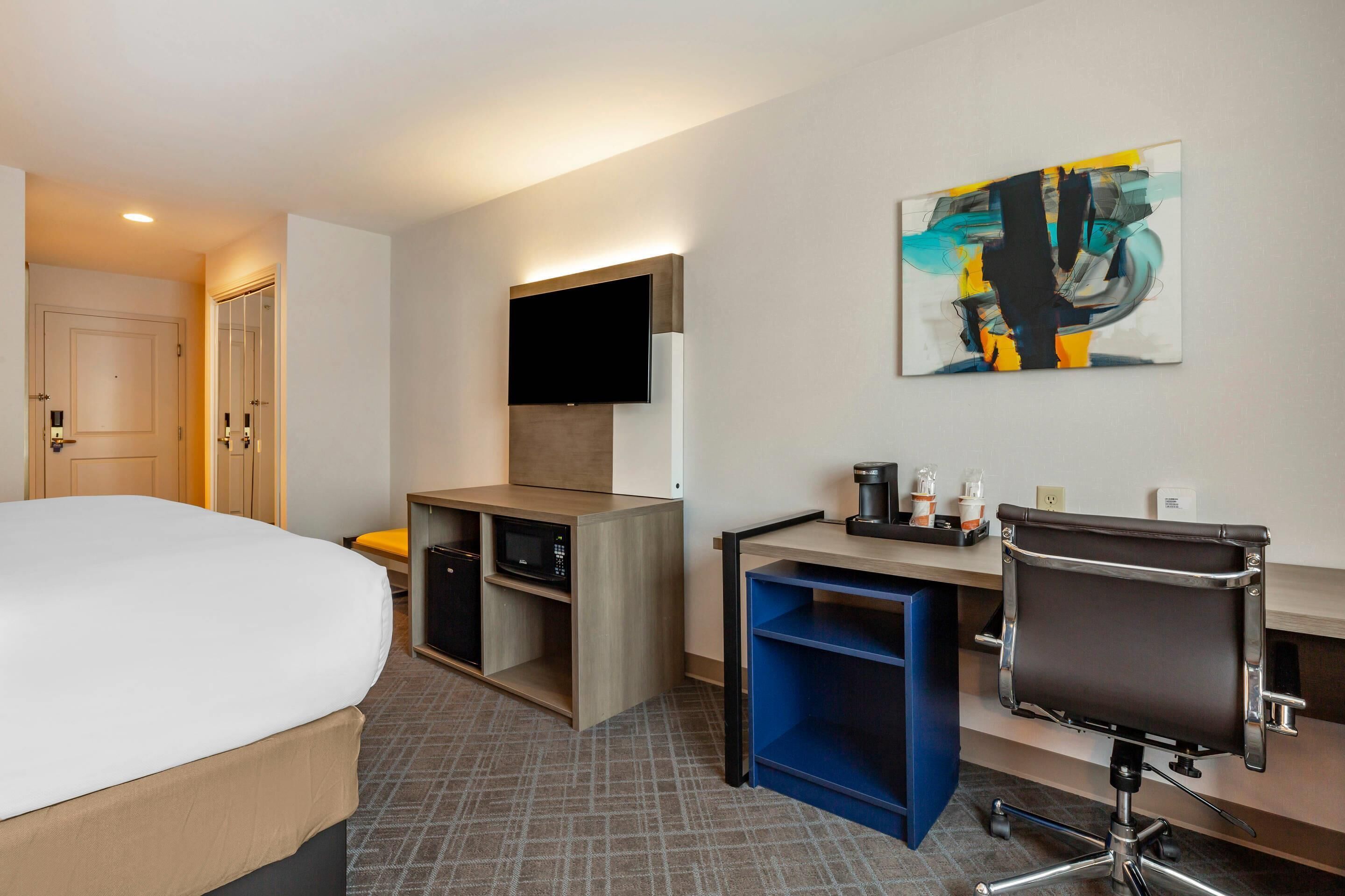 A hotel room with a bed, desk, chair, TV, and artwork.