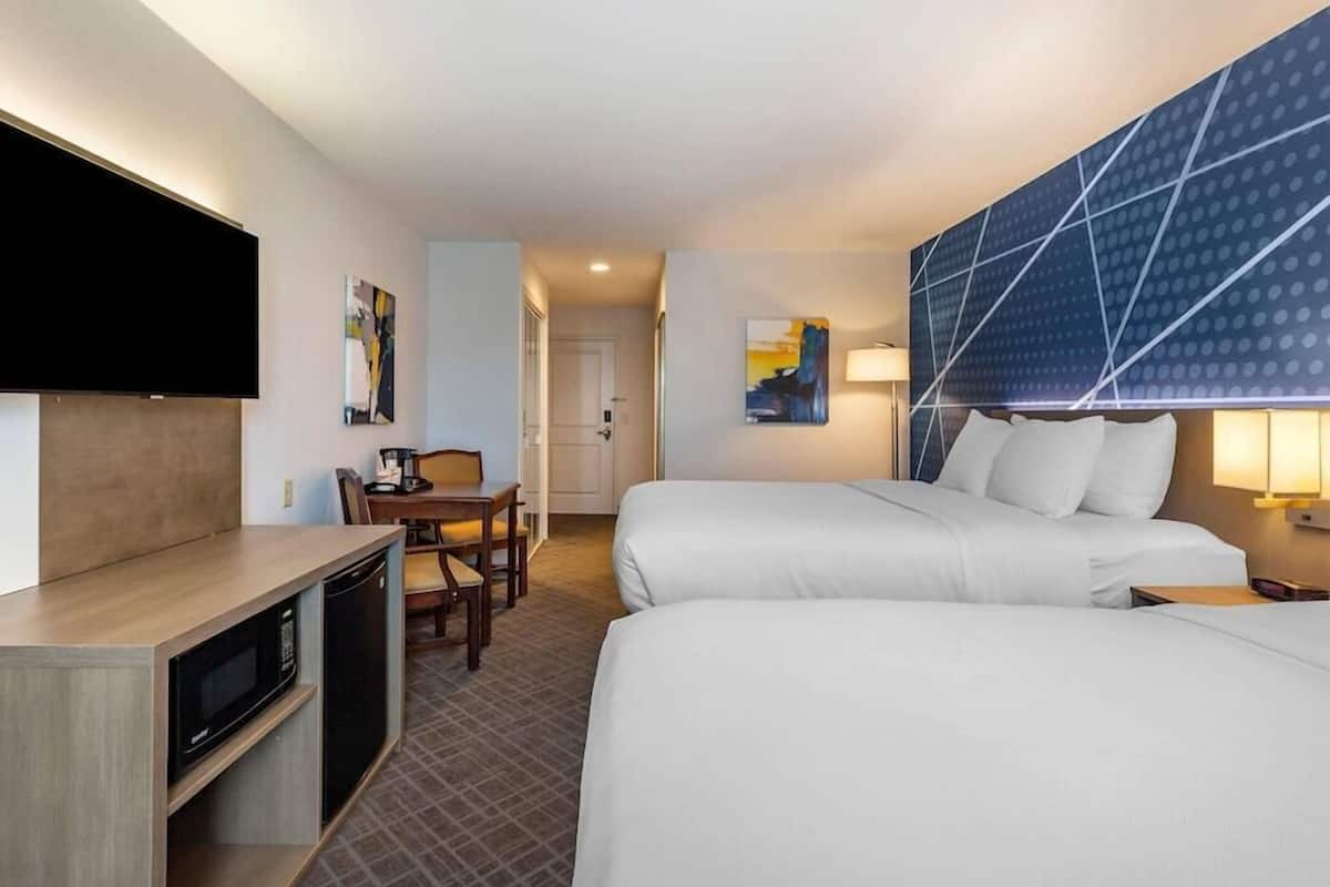 A hotel room with two beds, a desk with a chair, a flat-screen TV, a microwave, and a wall-mounted lamp.