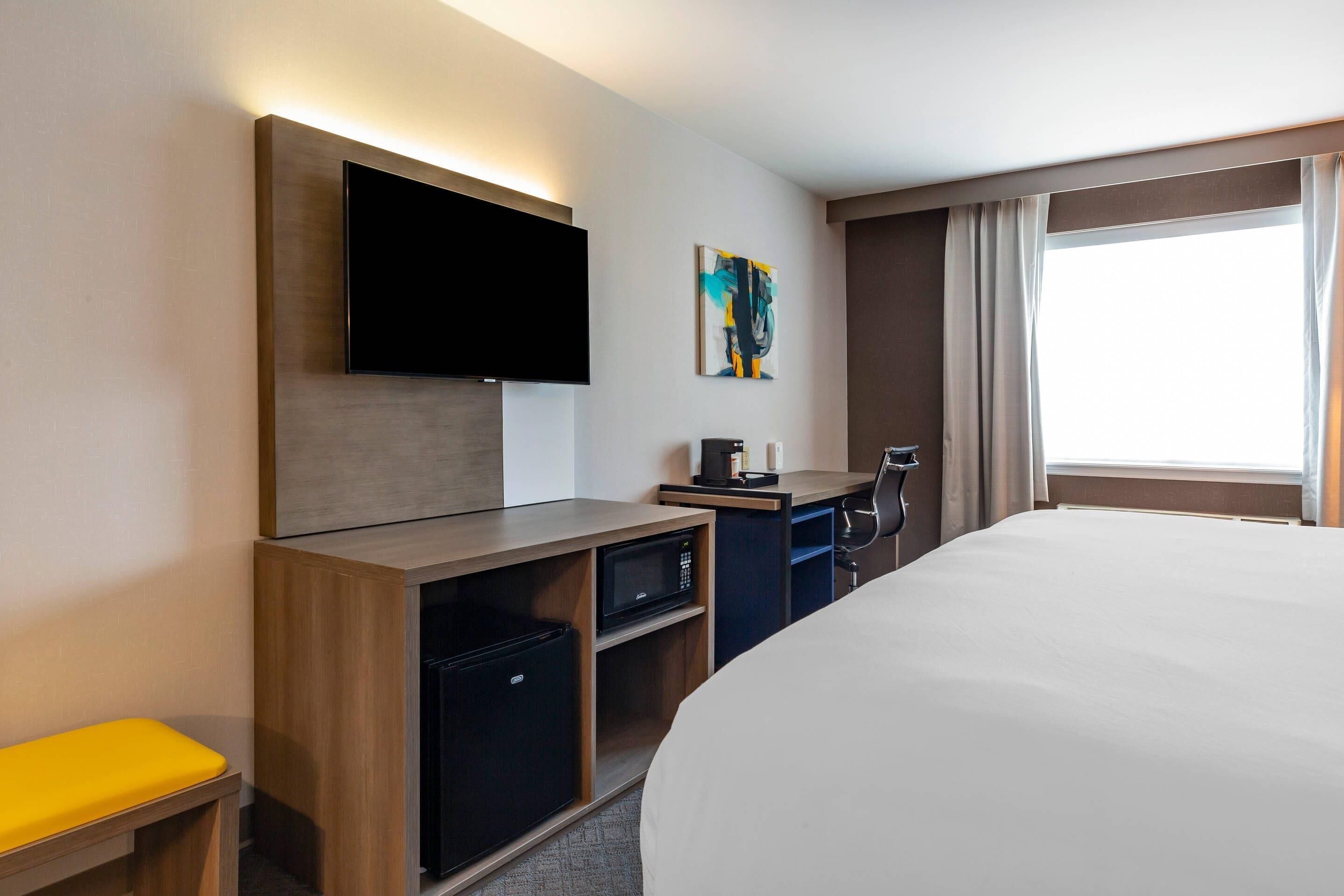 A hotel room with a bed, a flat-screen TV mounted on the wall, a desk with a computer, a microwave, and a small refrigerator.