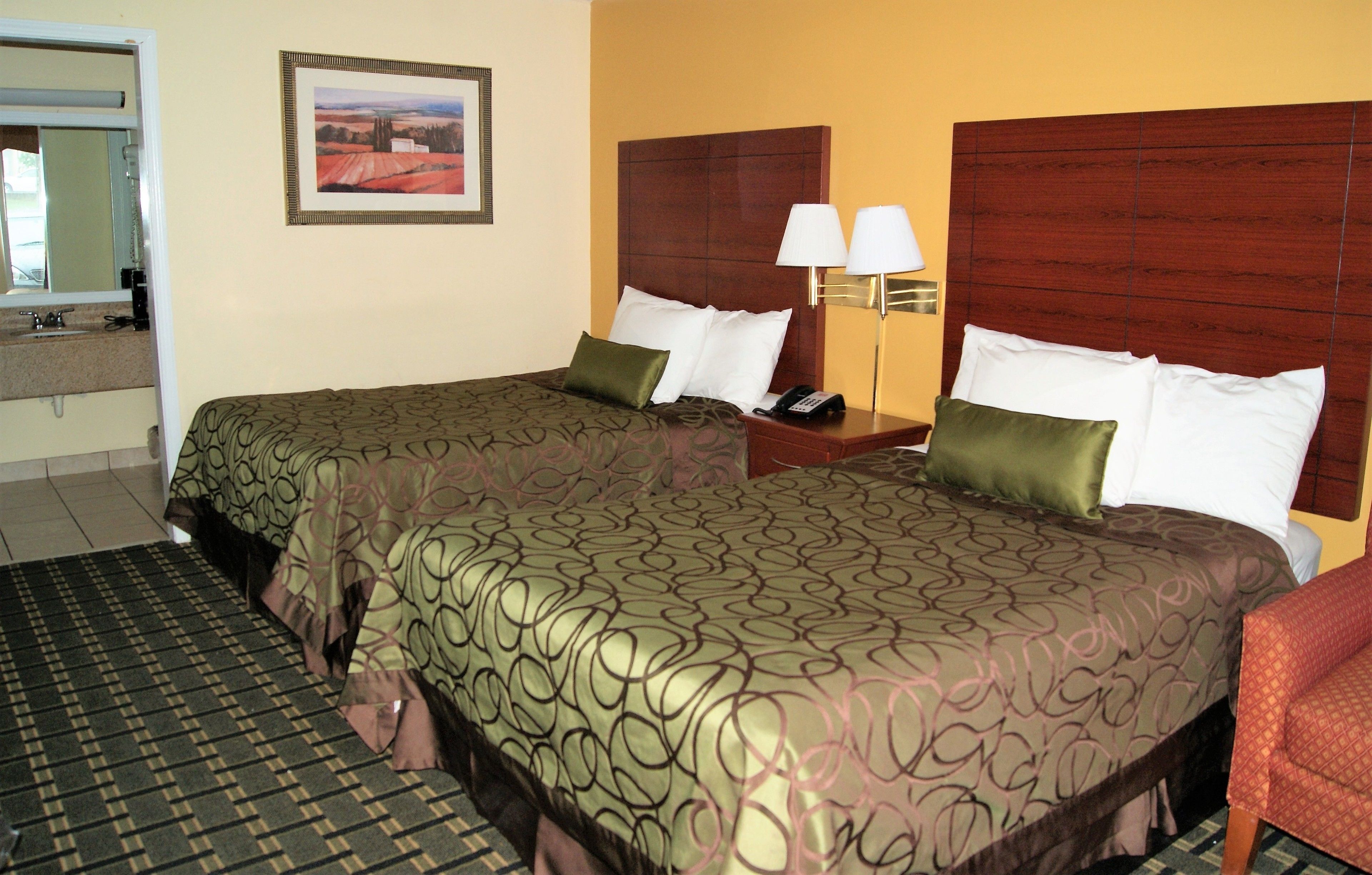 A hotel room with two beds, a nightstand with a lamp, a framed picture on the wall, and a bathroom visible through an open door.