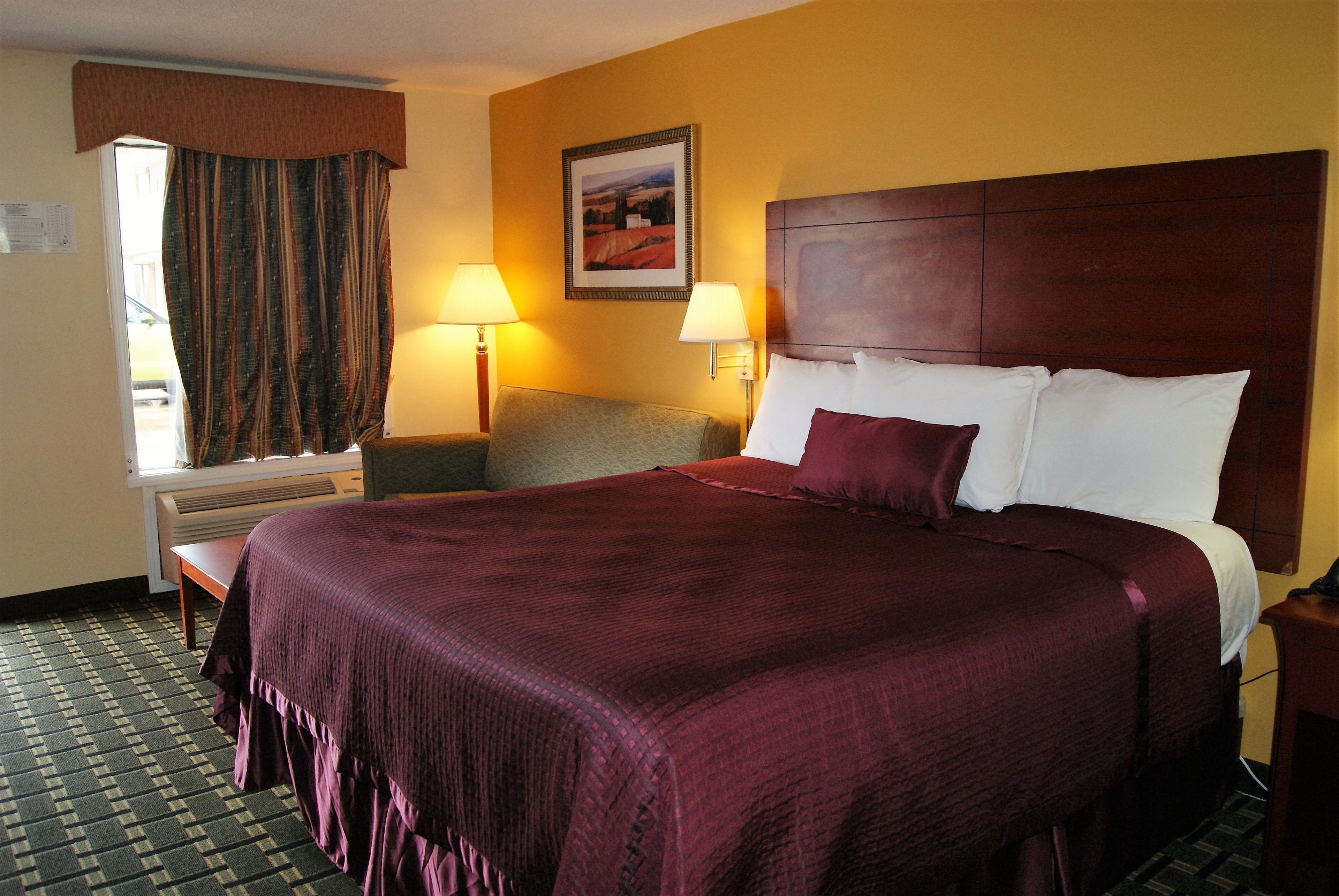 A hotel room with a large bed, two bedside lamps, a window with curtains, a small table, and a framed picture on the wall.