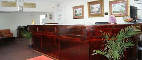 Un espace d’accueil avec un comptoir de réception en bois, une moquette rouge, des tableaux encadrés au mur et une plante en pot.