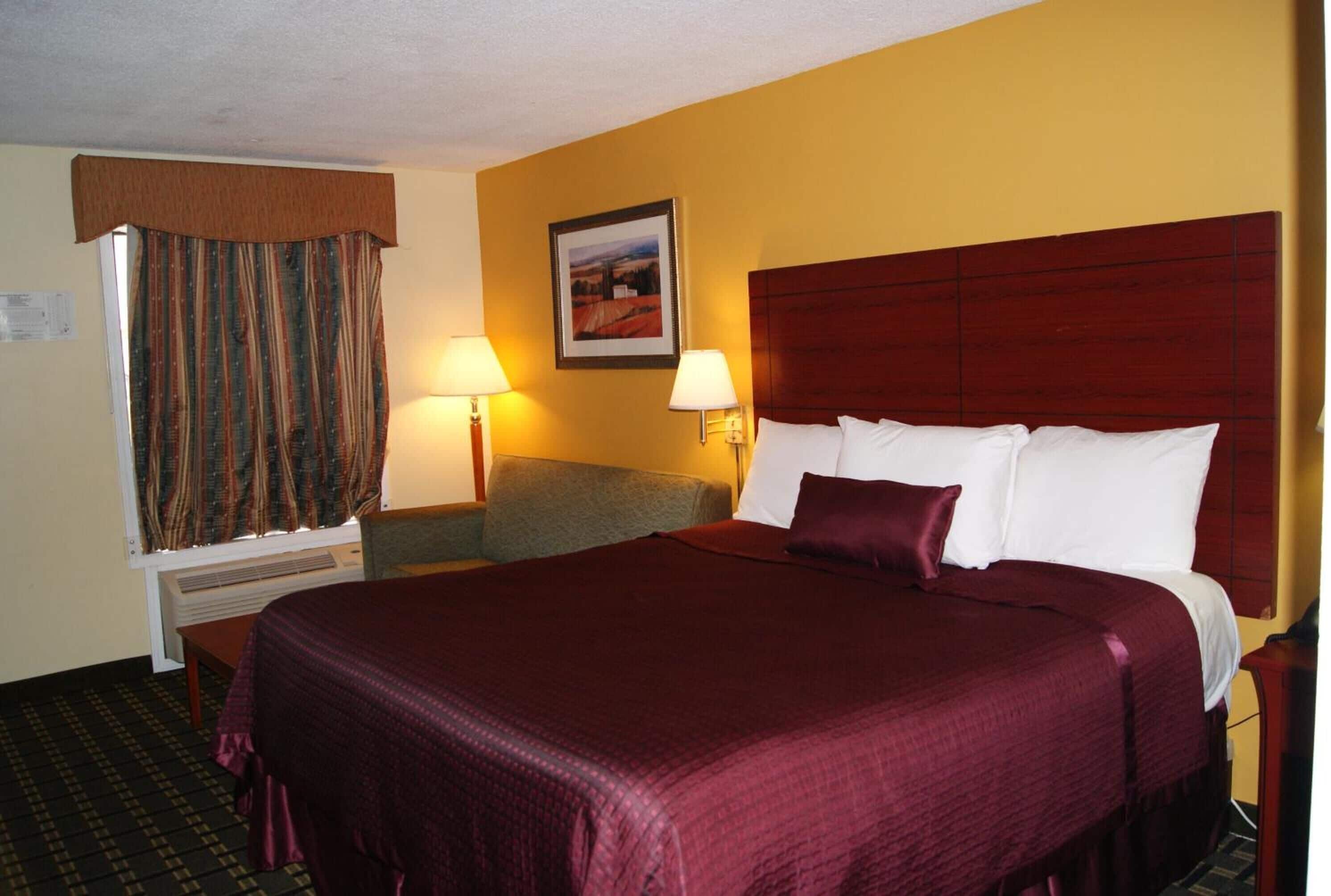 A hotel room with a large bed, a desk, and a chair. There is a window with curtains, a wall-mounted lamp, and a framed picture on the wall.