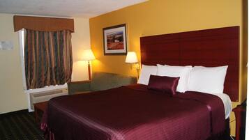 A hotel room with a large bed, a desk, and a chair. There is a window with curtains, a wall-mounted lamp, and a framed picture on the wall.