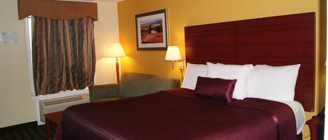 A hotel room with a large bed, a desk, and a chair. There is a window with curtains, a wall-mounted lamp, and a framed picture on the wall.