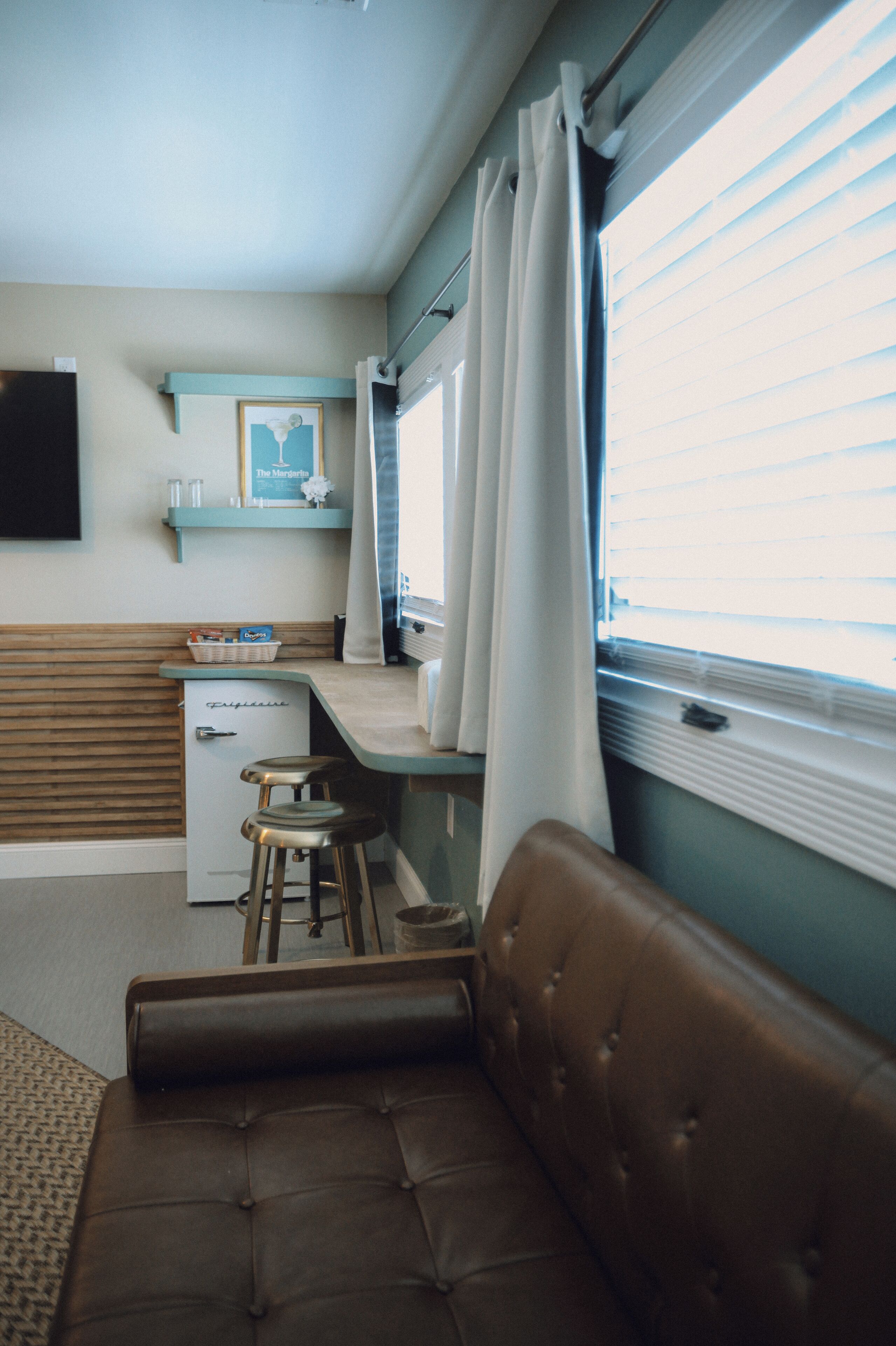 A compact kitchenette with a refrigerator, a small bar stool, and a window with blinds.