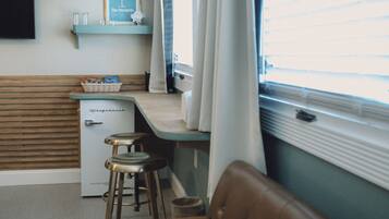A compact kitchenette with a refrigerator, a small bar stool, and a window with blinds.