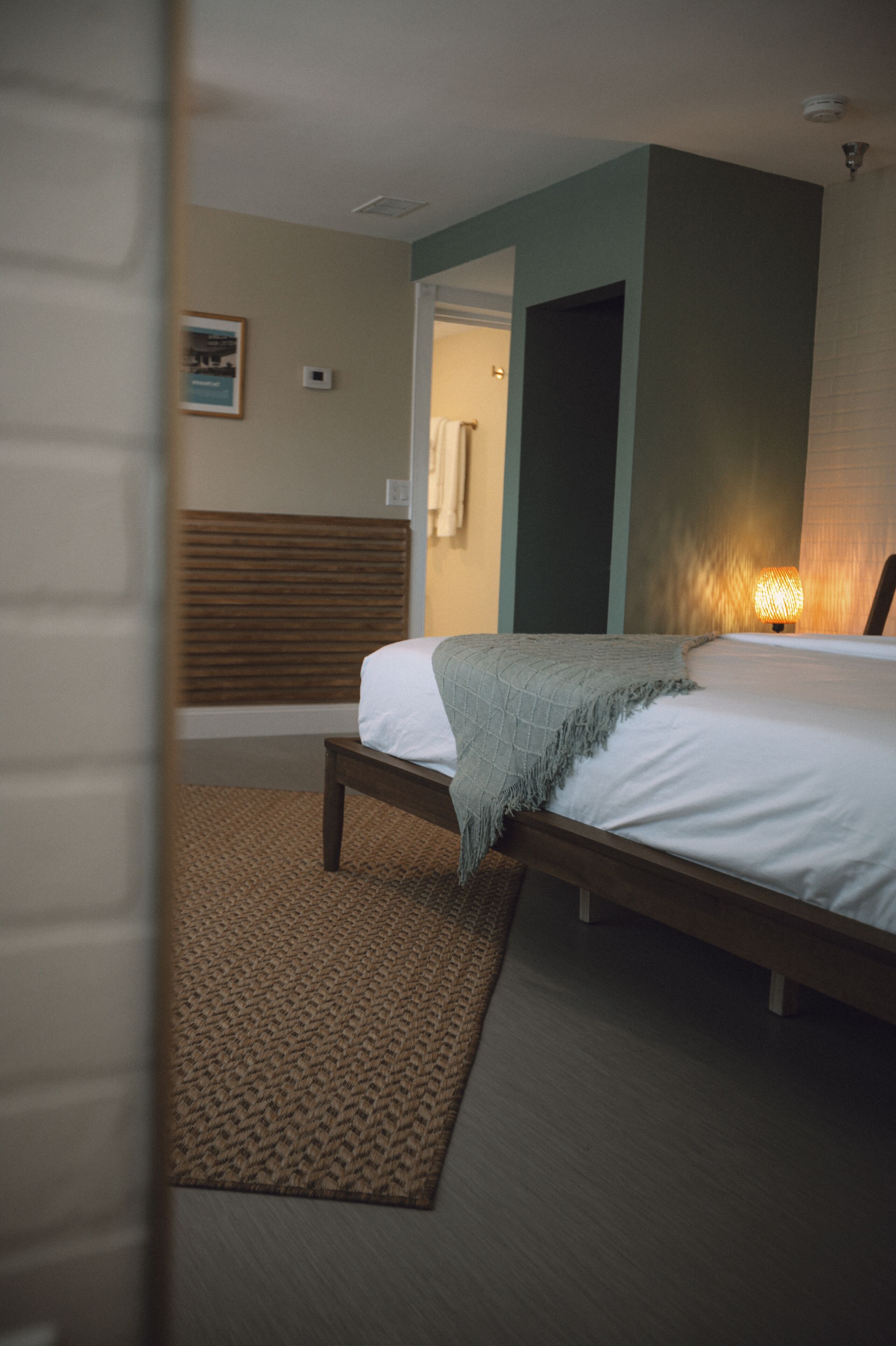 A bedroom with a bed, a bedside table, a wall-mounted lamp, and a door leading to another room.