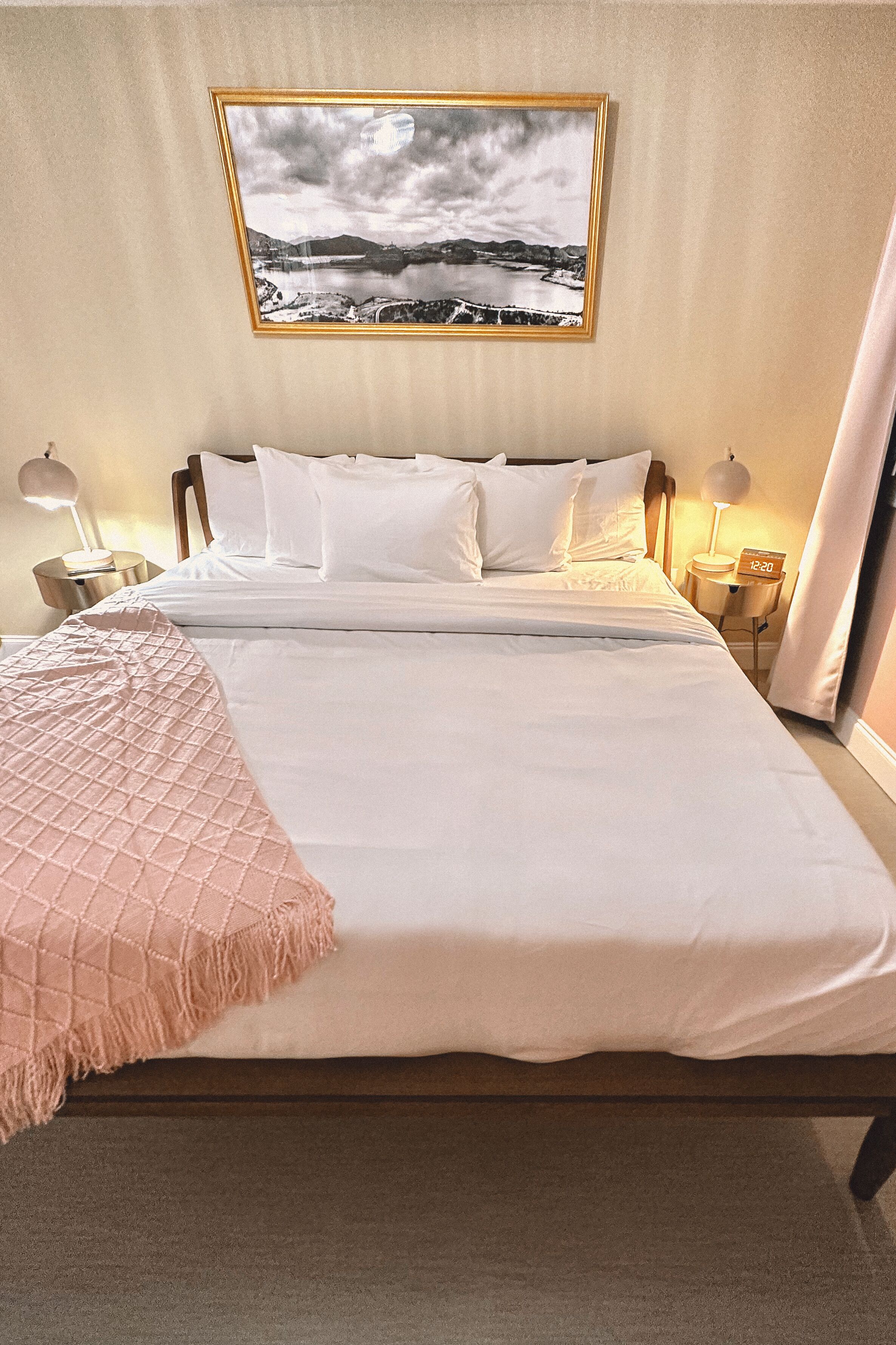 A neatly made bed with white linens and a pink throw blanket, two bedside lamps, a framed landscape picture on the wall, and a clock on the nightstand.