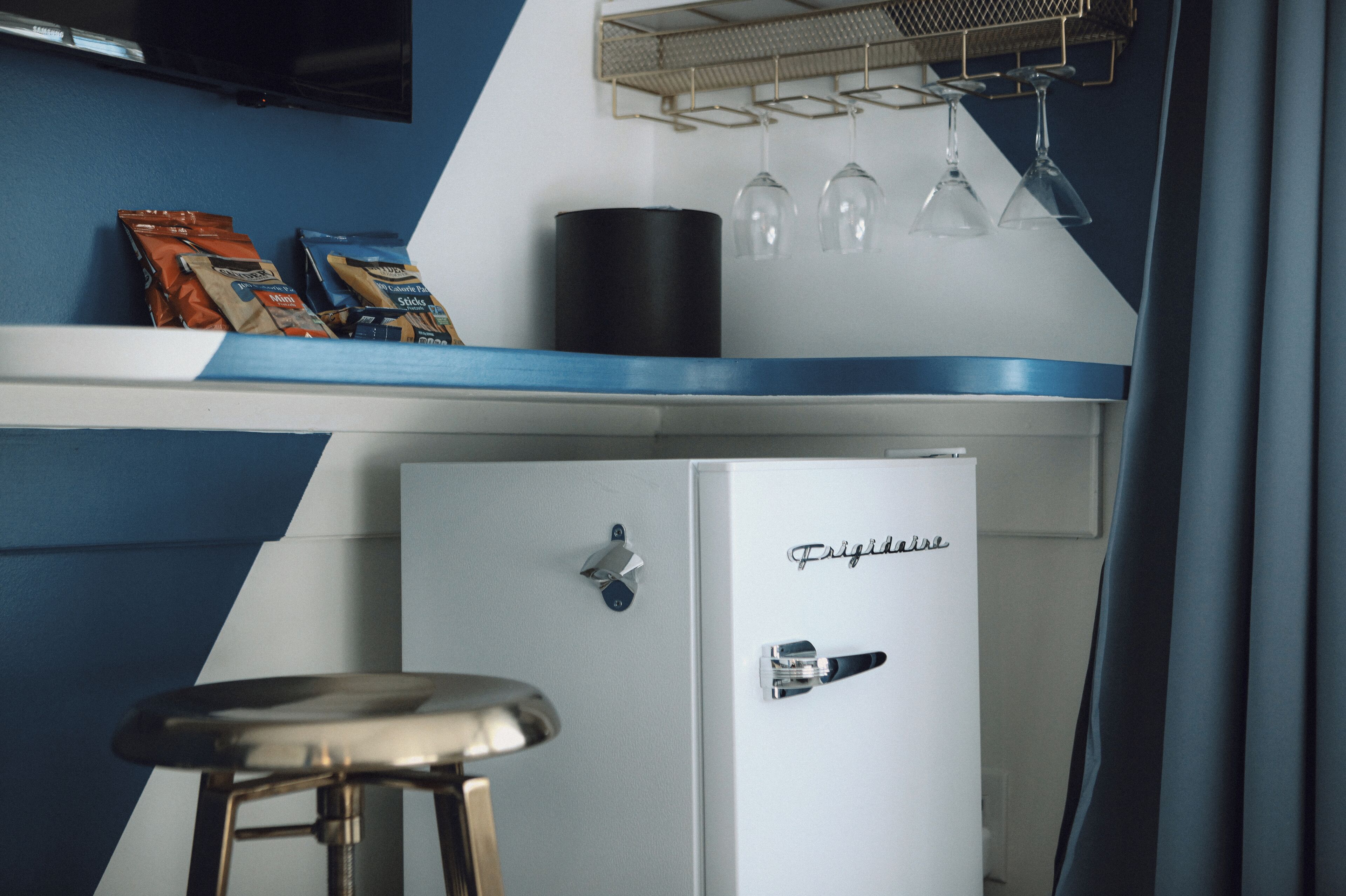 A compact kitchen area with a refrigerator, a bar stool, and a shelf with snacks and wine glasses.