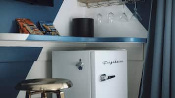 A compact kitchen area with a refrigerator, a bar stool, and a shelf with snacks and wine glasses.