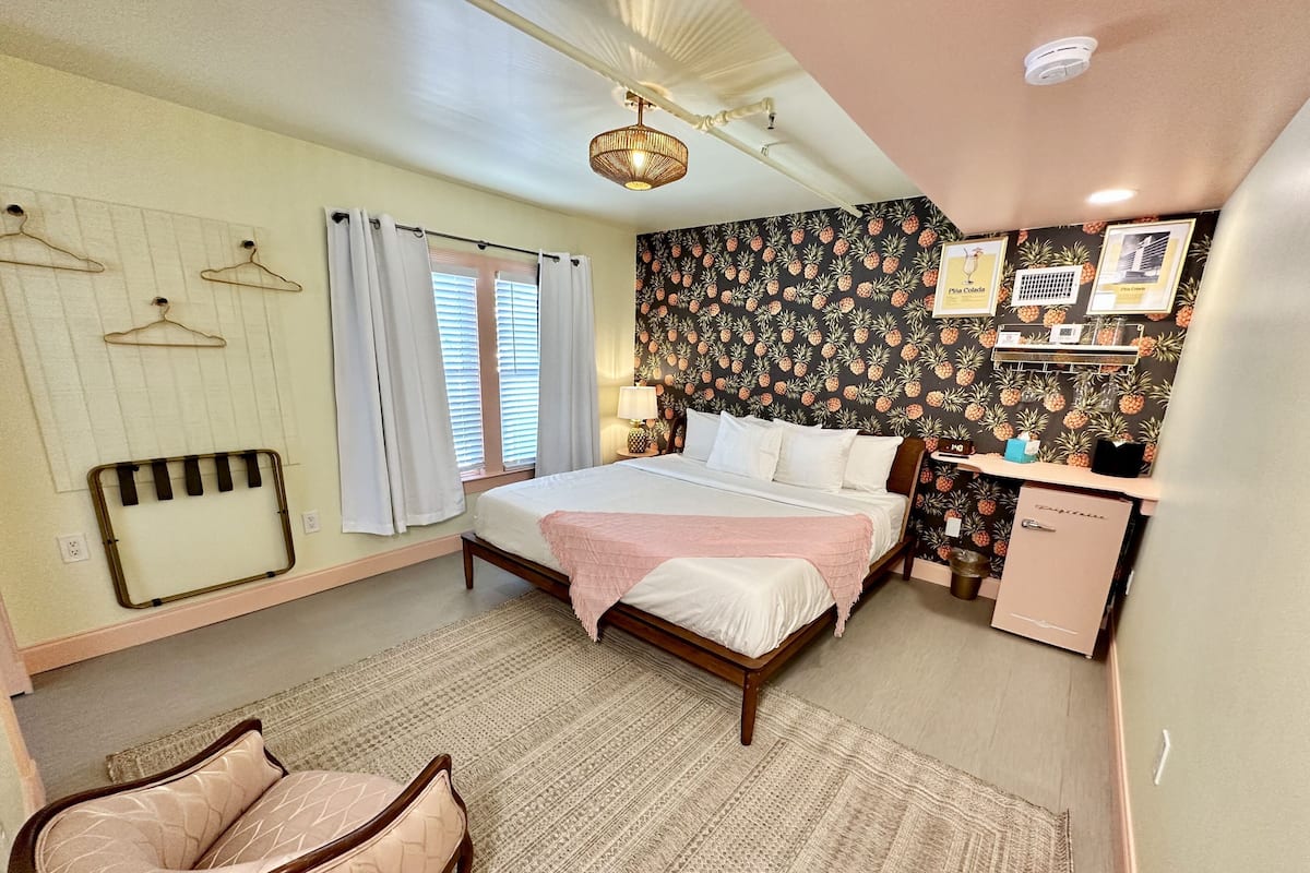 A bedroom with a bed, bedside table, lamp, and a small kitchenette.