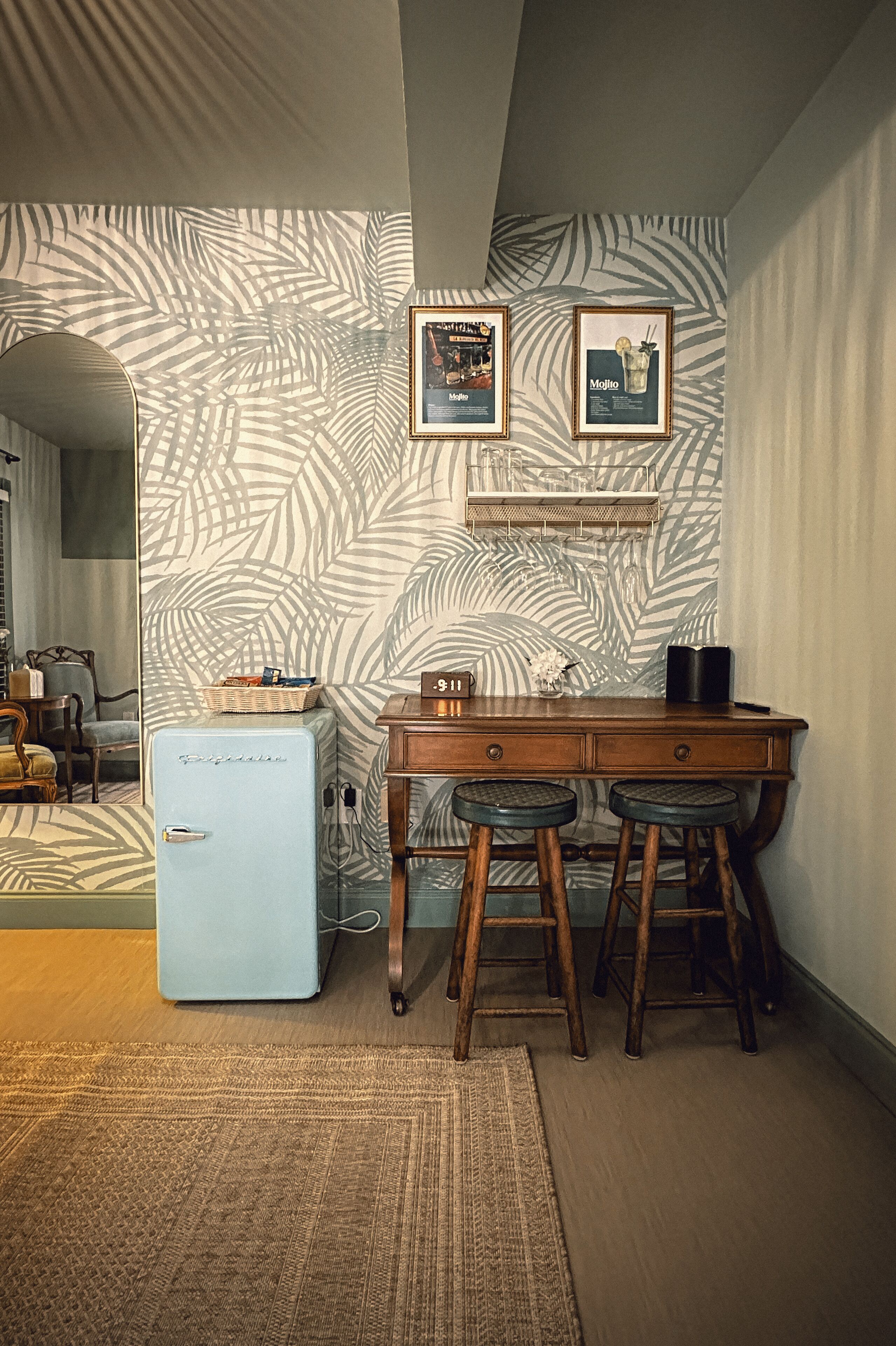 Una habitación pequeña con un refrigerador de estilo vintage, un escritorio de madera con dos taburetes y un papel tapiz estampado.