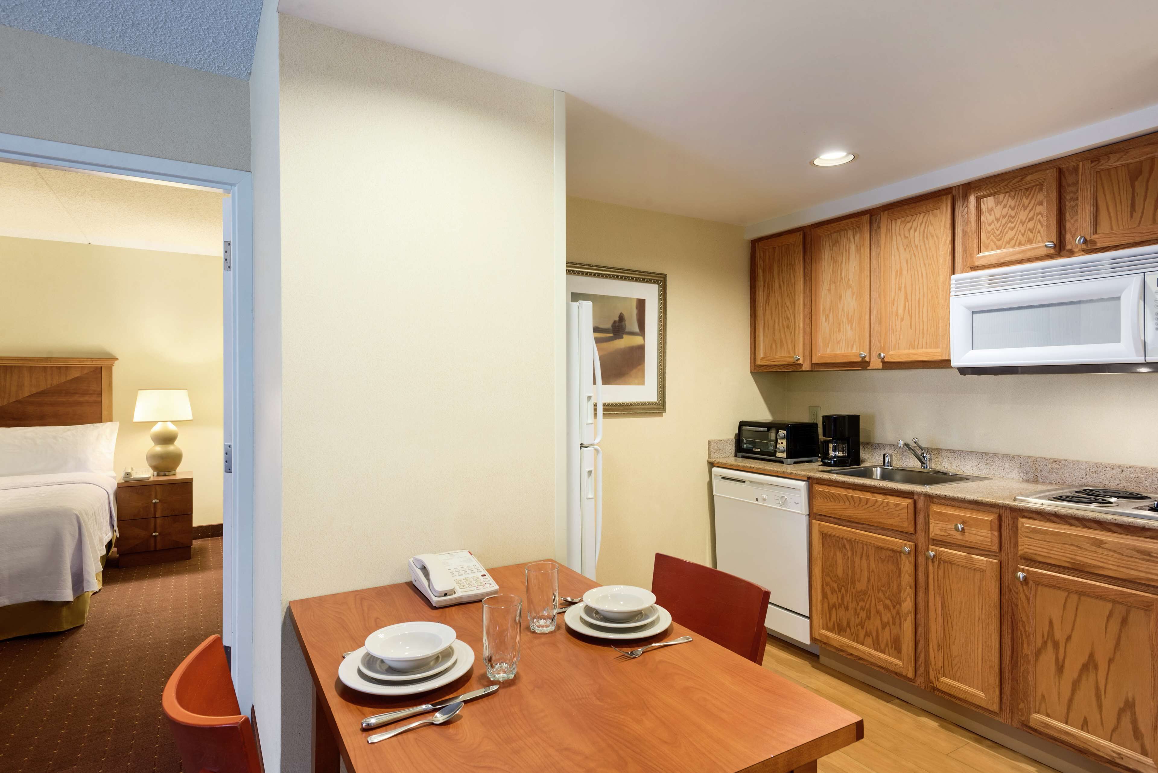 A hotel room with a wooden dining table set for two, a bed, a desk with a lamp, and a kitchenette with wooden cabinets and a microwave.