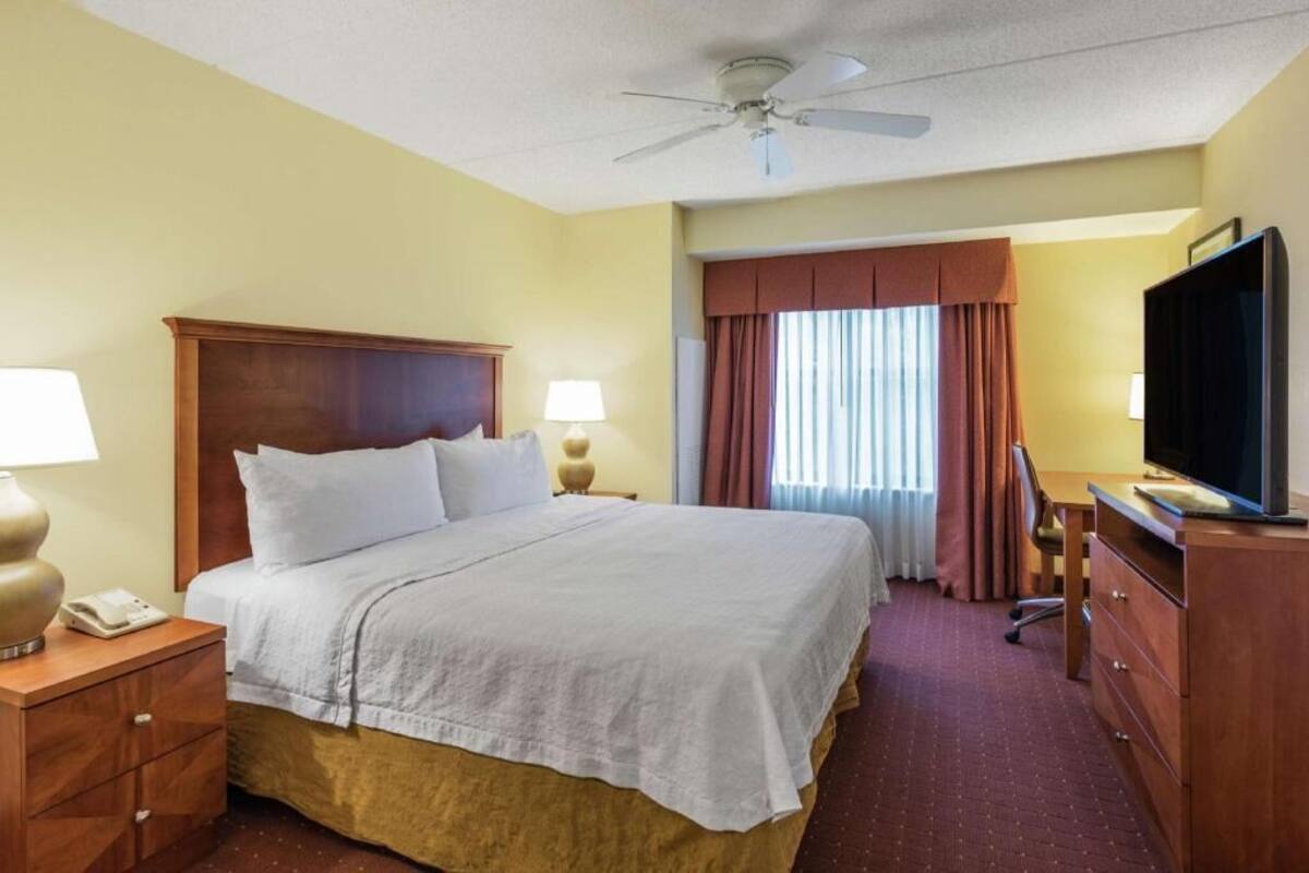 A hotel room with a bed, bedside lamps, a television, a desk, and a ceiling fan.