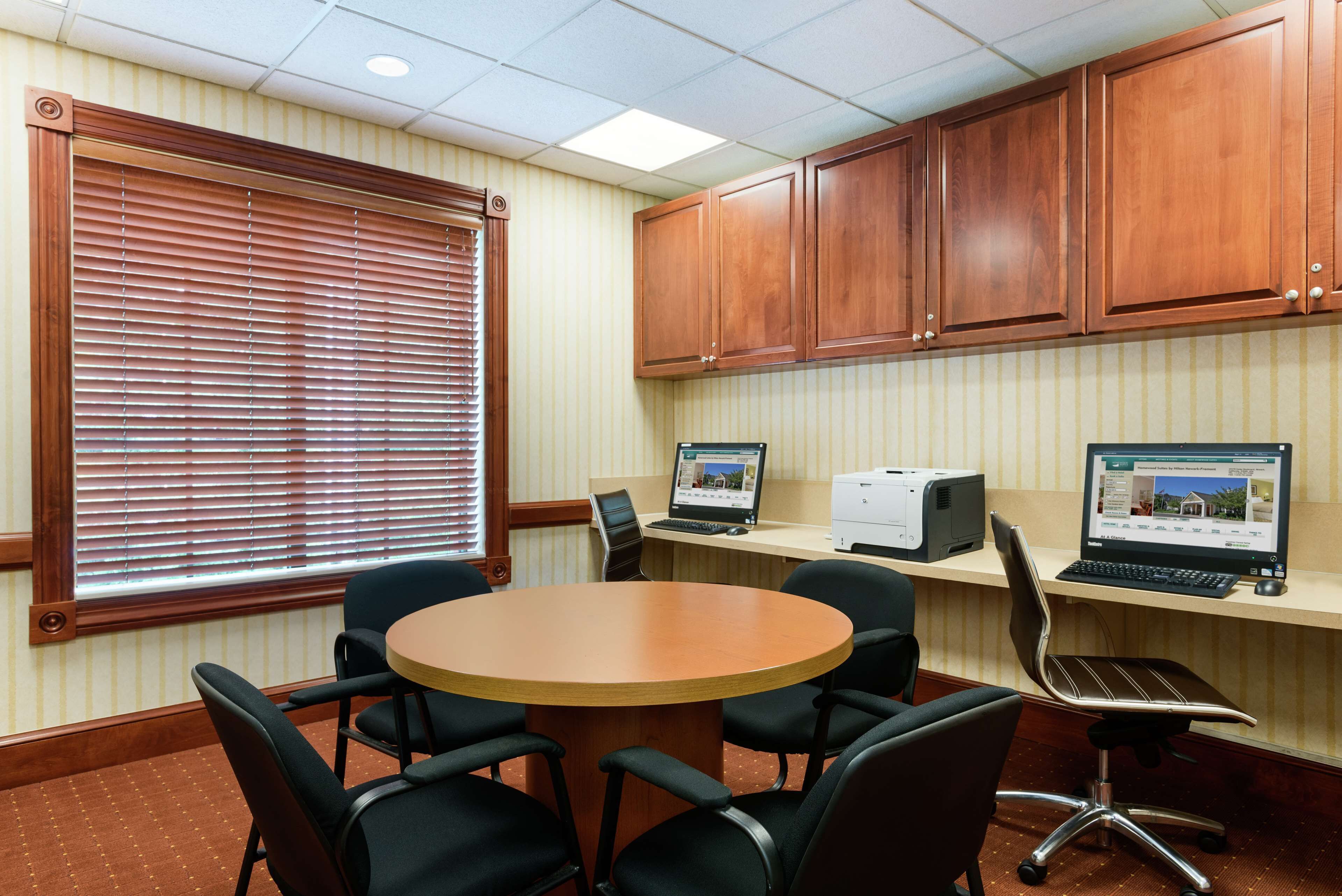 A conference room with a round table, six chairs, wooden cabinets, two desktop computers, and a window with blinds.