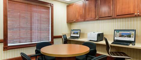 A conference room with a round table, six chairs, wooden cabinets, two desktop computers, and a window with blinds.