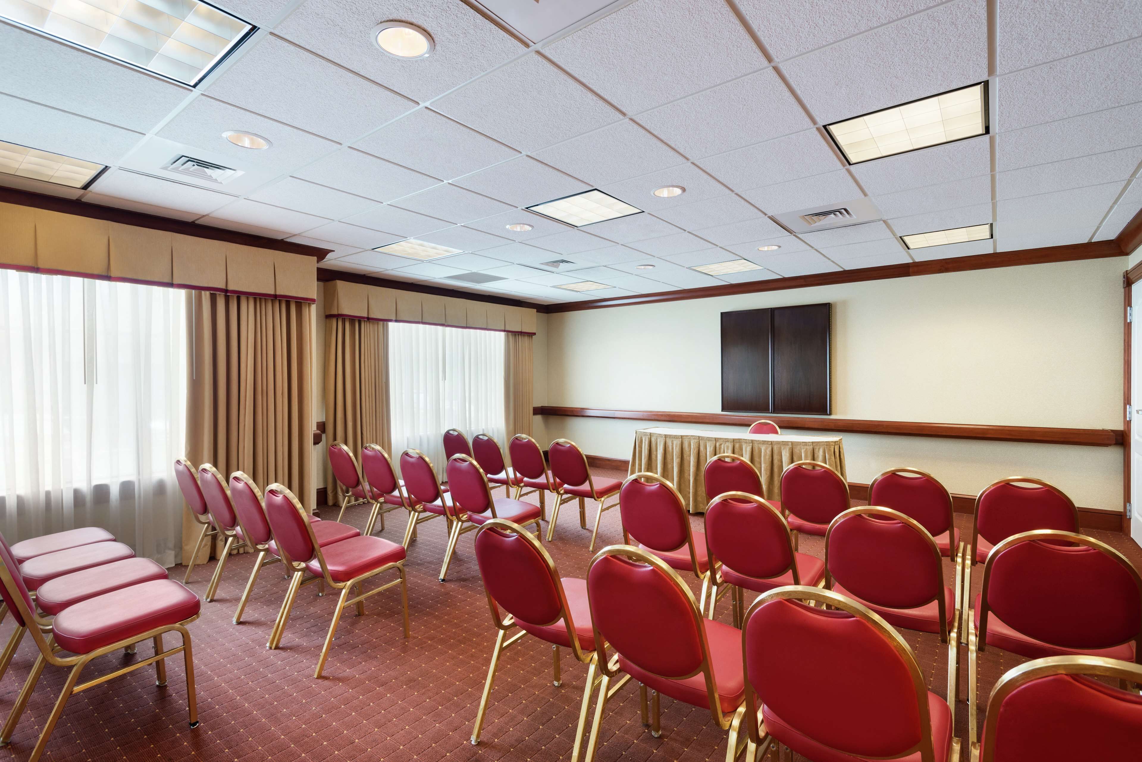 A conference room with rows of red chairs, a large screen, and a window with curtains.
