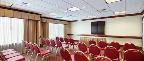 A conference room with rows of red chairs, a large screen, and a window with curtains.