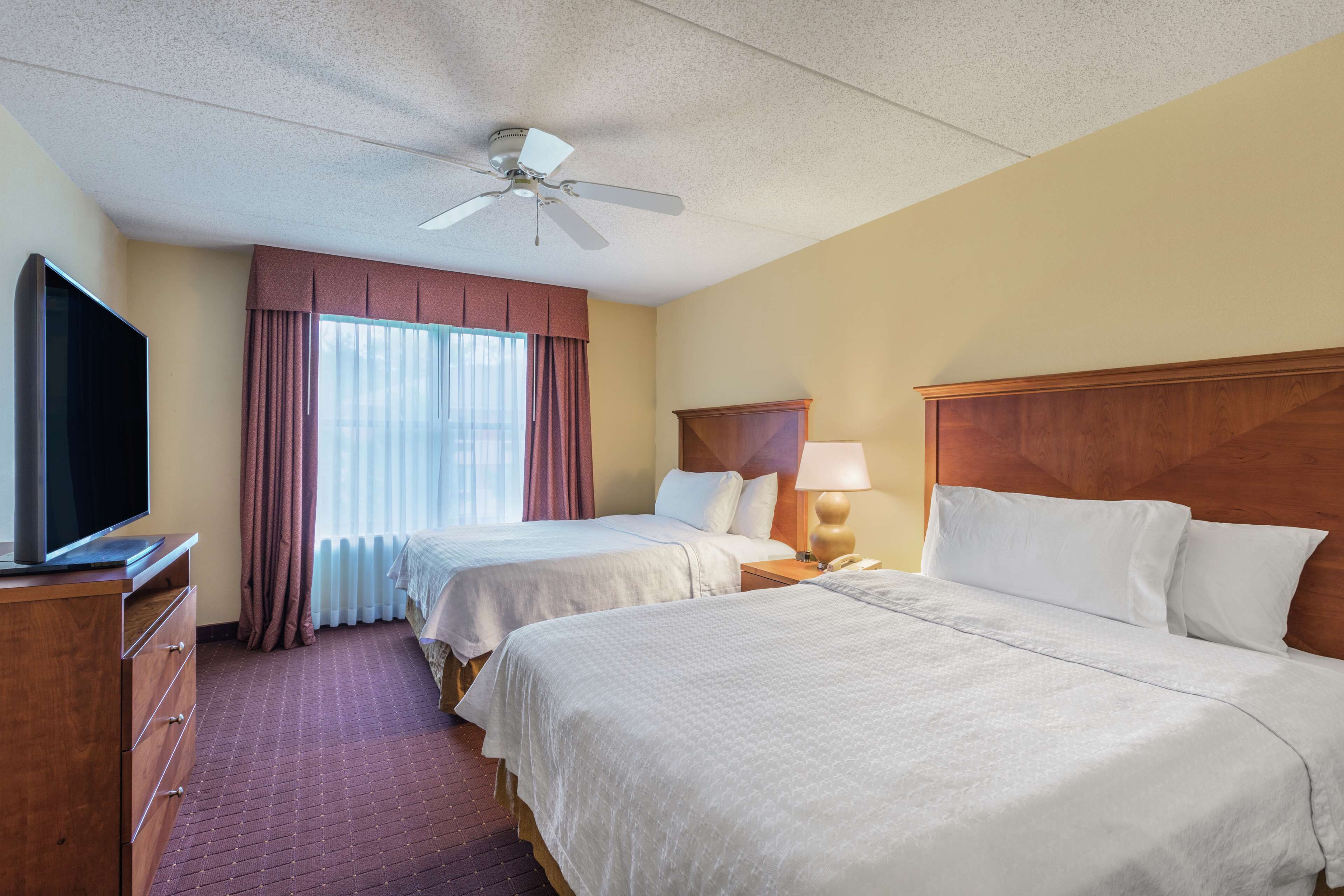 A hotel room with two beds, a TV, a ceiling fan, and a window with curtains.
