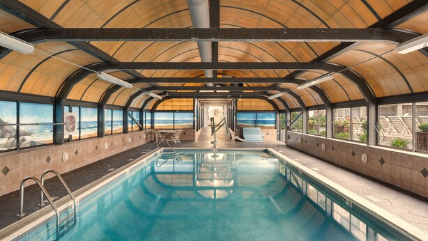 Una piscina interior con vistas a la playa a través de amplias ventanas, suelo de baldosas y una estructura de techo curva.