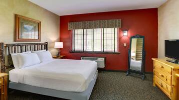 A hotel room with a large bed, a wooden dresser, a TV, a window with blinds, a wall-mounted lamp, and a framed picture on the wall.