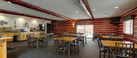 A large dining area with wooden tables and chairs, a kitchen area with a trash bin, and a television on the wall.