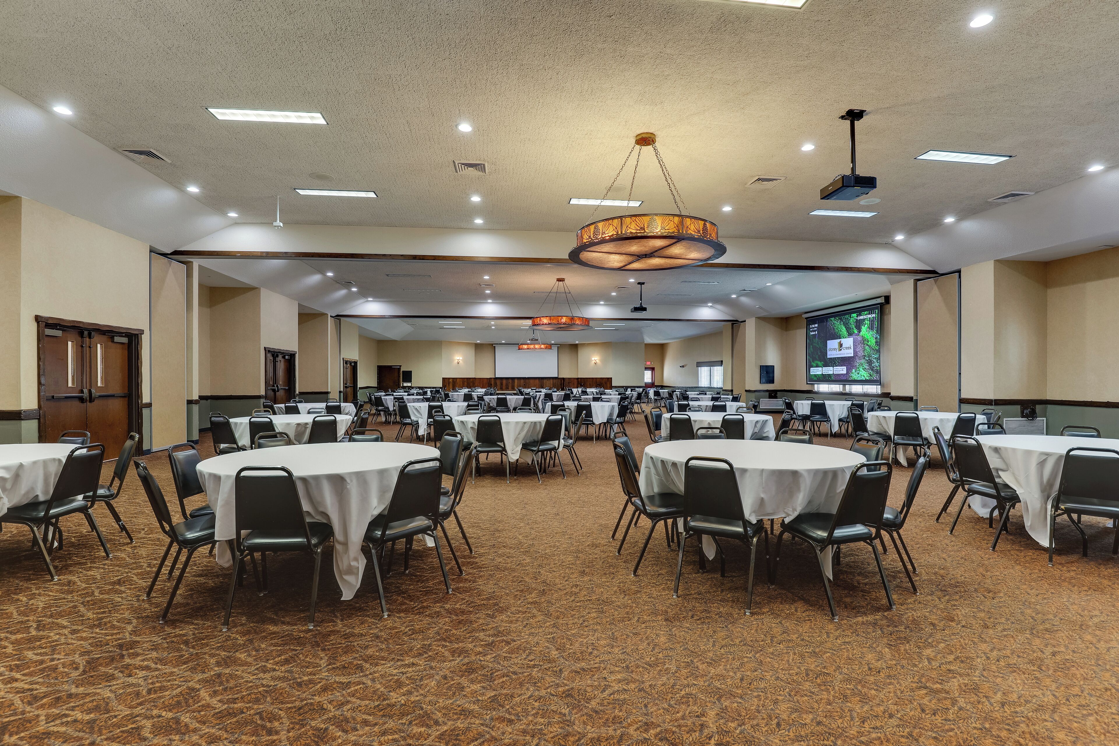 A large conference room with multiple round tables and chairs, carpeted flooring, and a ceiling with recessed lighting and a central chandelier.