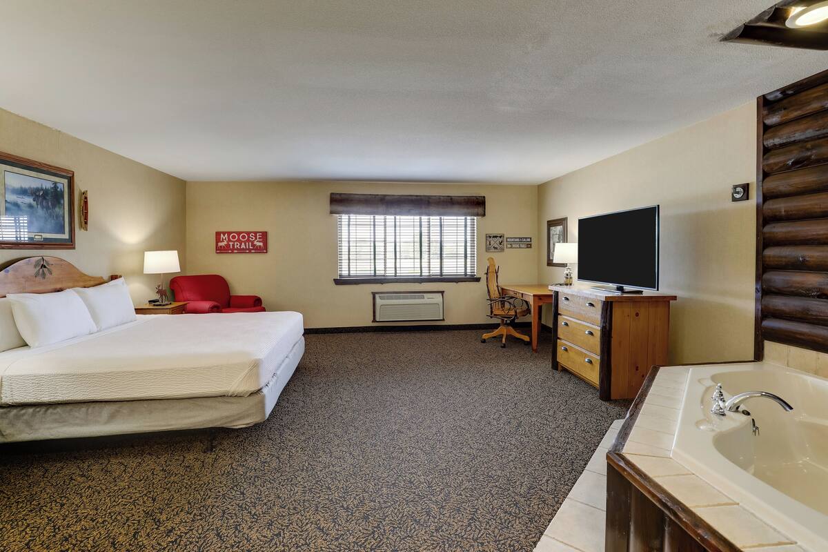 A hotel room with a bed, a desk with a chair, a television, a large window with blinds, a wooden wall panel, and a bathroom with a bathtub.