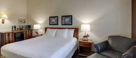 Una habitación de hotel con una cama grande, dos mesitas de noche con lámparas, un escritorio con teléfono y cafetera, y un sillón de cuero.
