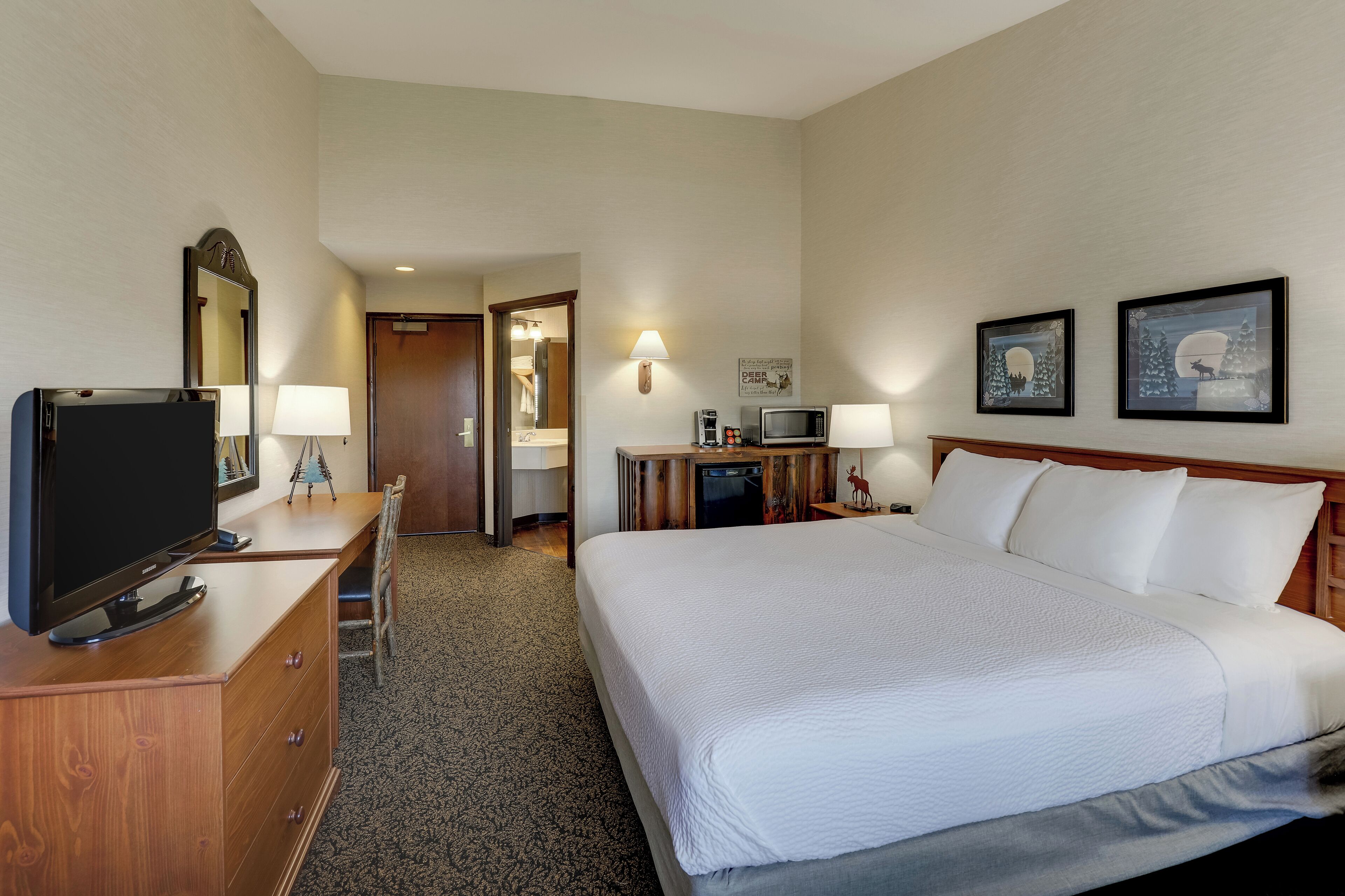 A hotel room with a large bed, a TV on a wooden dresser, a desk with a lamp, and a microwave.