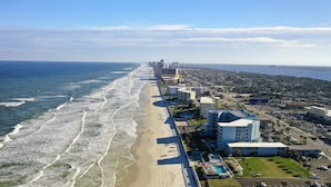 Vista aérea de una ciudad costera con un hotel frente a la playa, que incluye una zona de piscina y edificios adyacentes.