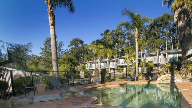A swimming pool with a brick border, surrounded by palm trees and a fence. - Fairway Motor Inn (Pambula)