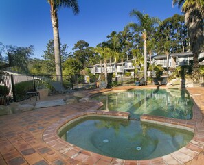 A swimming pool with a brick border, surrounded by palm trees and a fence. - Fairway Motor Inn (Pambula)