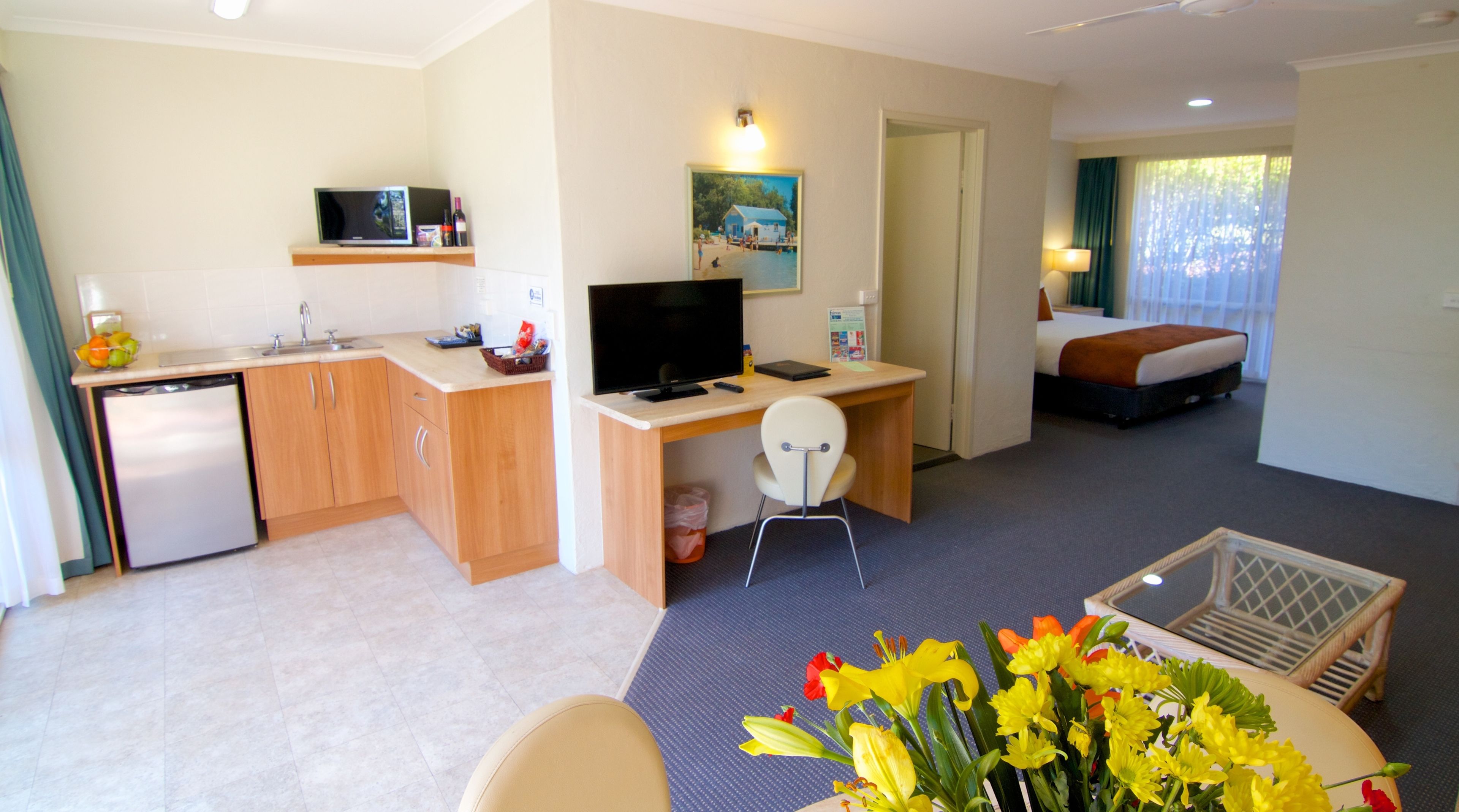 A compact hotel room with a kitchenette, a desk with a computer, a bed, and a small seating area with flowers.