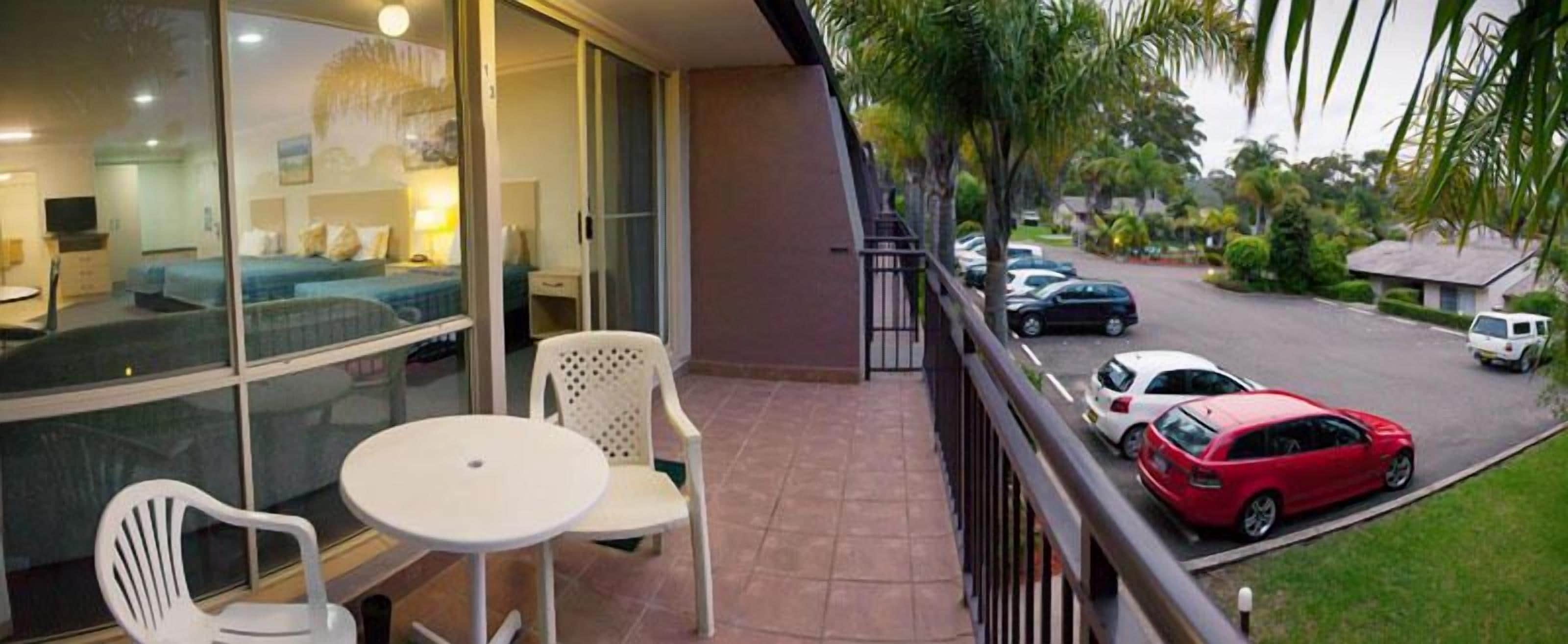 Un balcón con mesa y sillas, vista a una calle con coches aparcados y un vistazo a una habitación de hotel con cama y televisor.