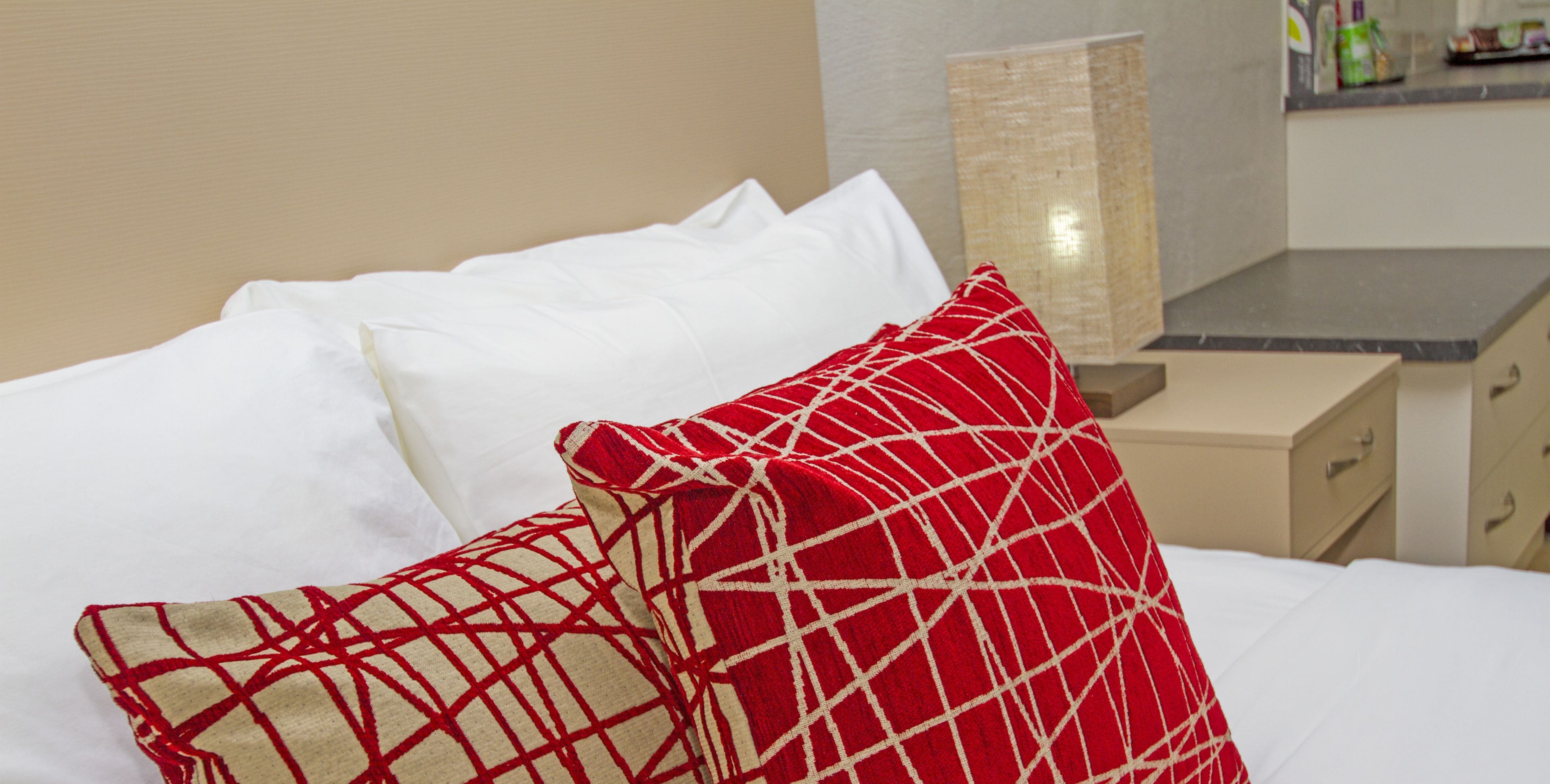 Une chambre d’hôtel avec un lit, des oreillers blancs et rouges à motifs, une table de chevet beige et un miroir.