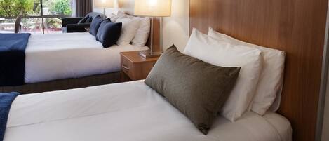 Habitación de hotel con dos camas, cada una con sábanas blancas y numerosas almohadas. Hay mesitas de noche con lámparas y vistas al verdor exterior.