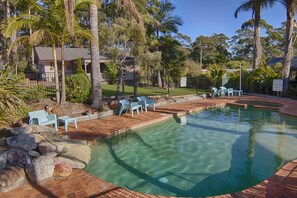 A clear swimming pool with a brick deck, surrounded by palm trees and a fenced area with chairs.