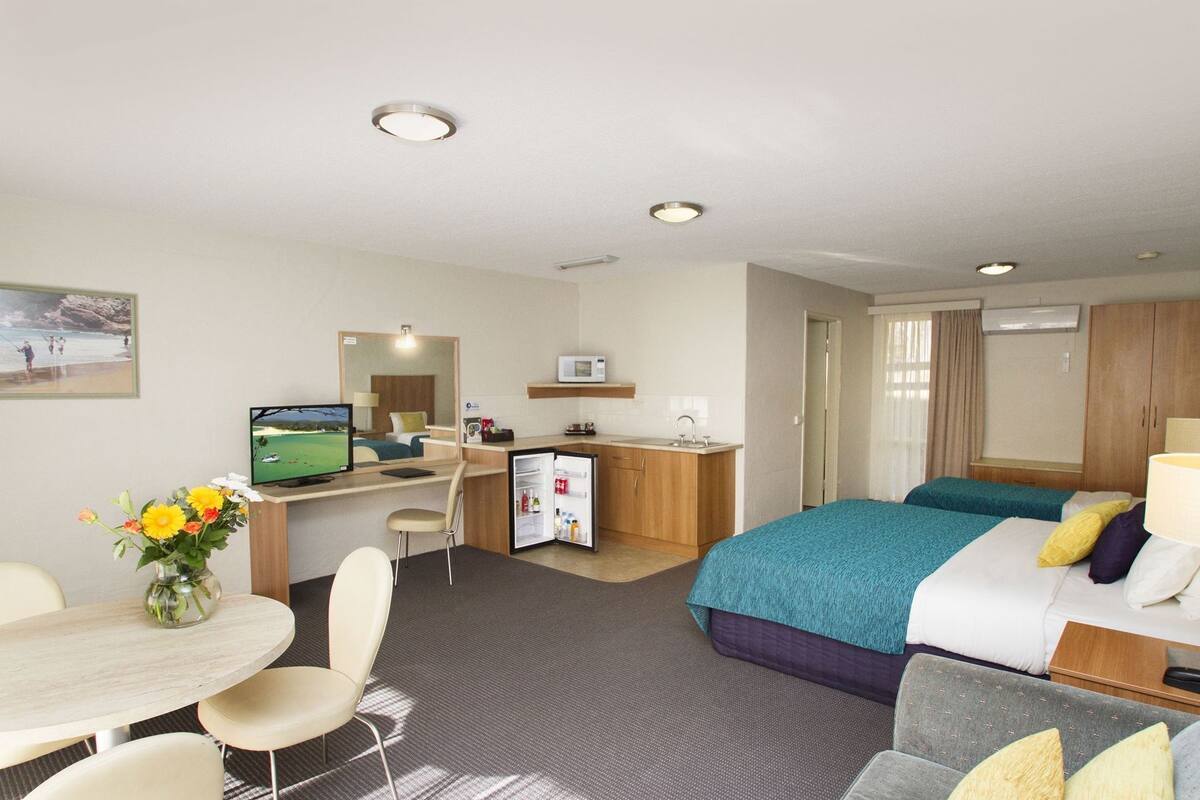 A hotel room with a bed, a desk with a computer, a mini-fridge, a TV, and a dining area with a table and chairs.