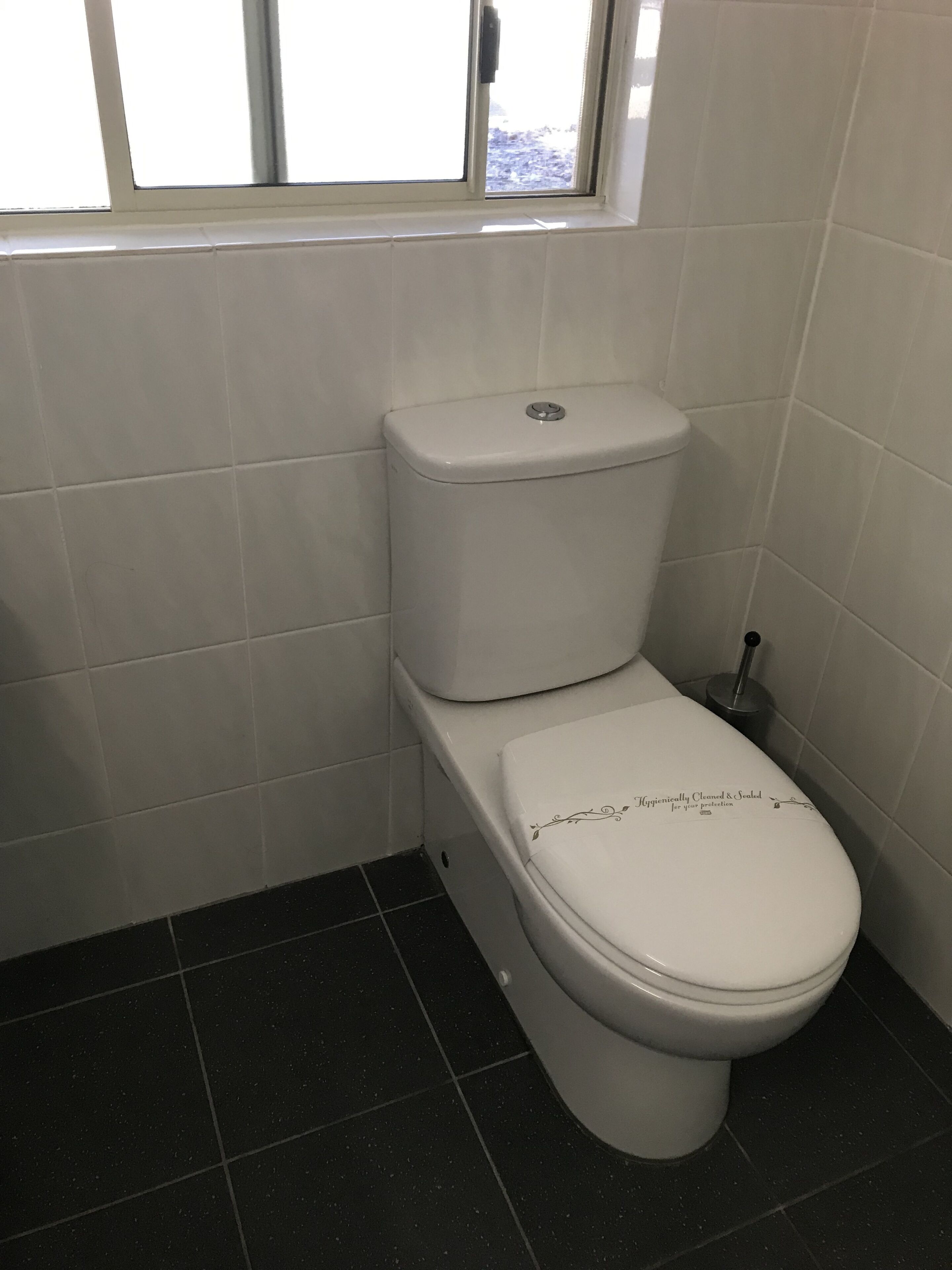 A standard toilet with a white seat and lid, a flush handle, and a window above the toilet.